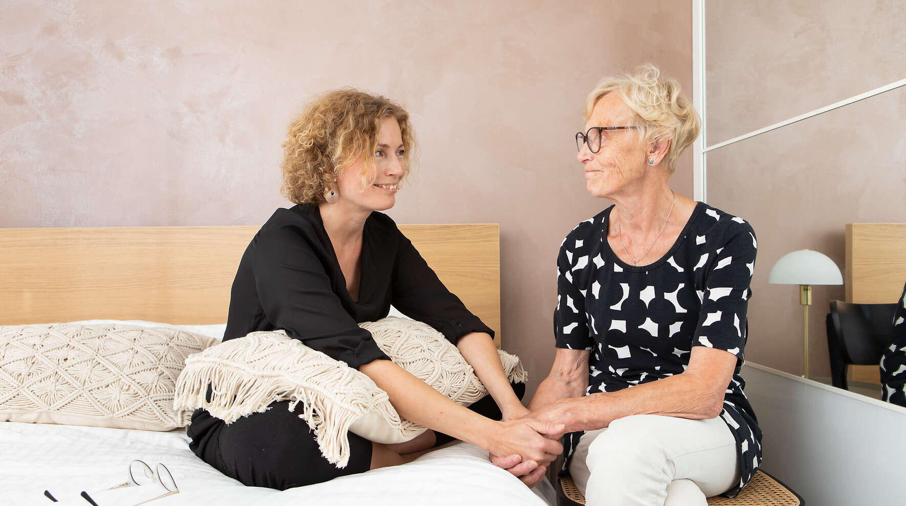Keski-ikäinen nainen ja iäkäs nainen istuvat kädet yhteen liitettyinä ja katsovat toisiaan hymyillen.