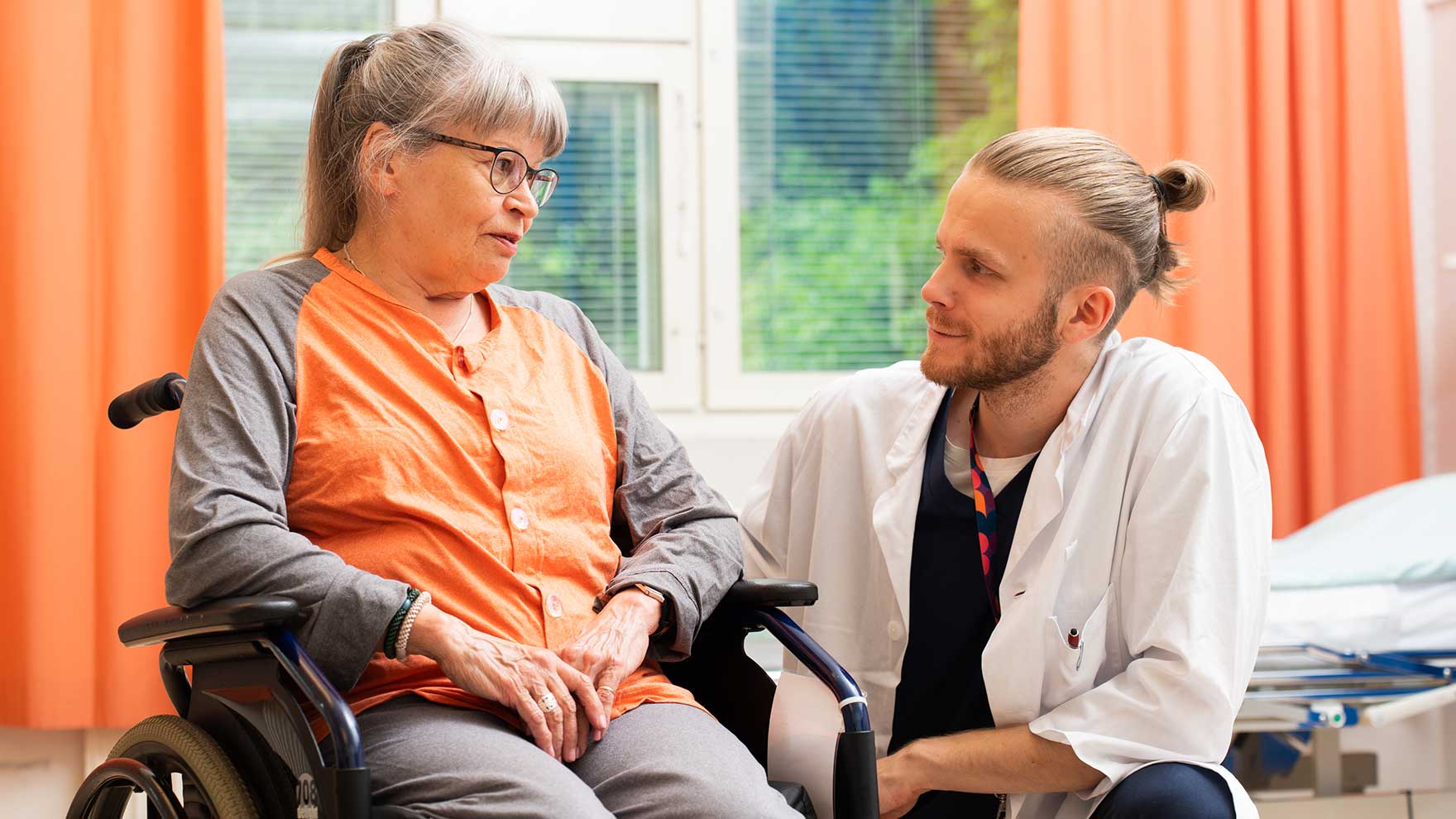 Pyörätuolissa istuva naispotilas juttelee lääkärin kanssa sairaalaosastolla.