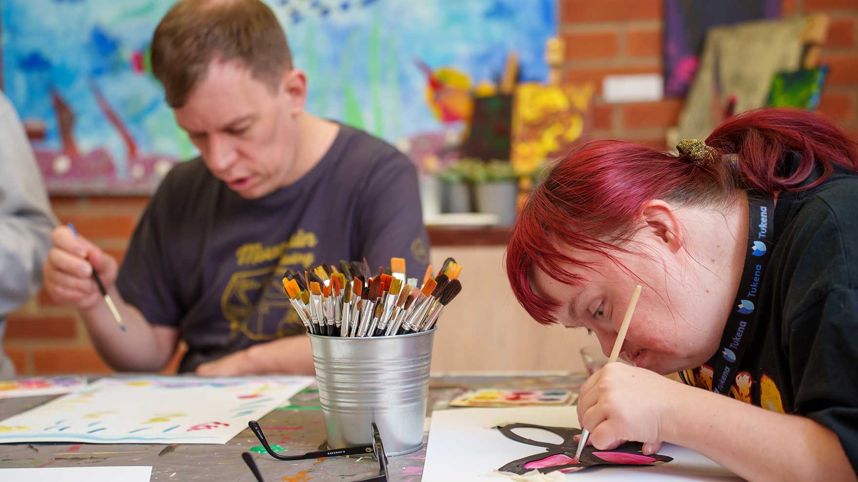 A woman and a man are painting intently with watercolors. In the middle of the table is a jar of paintbrushes.