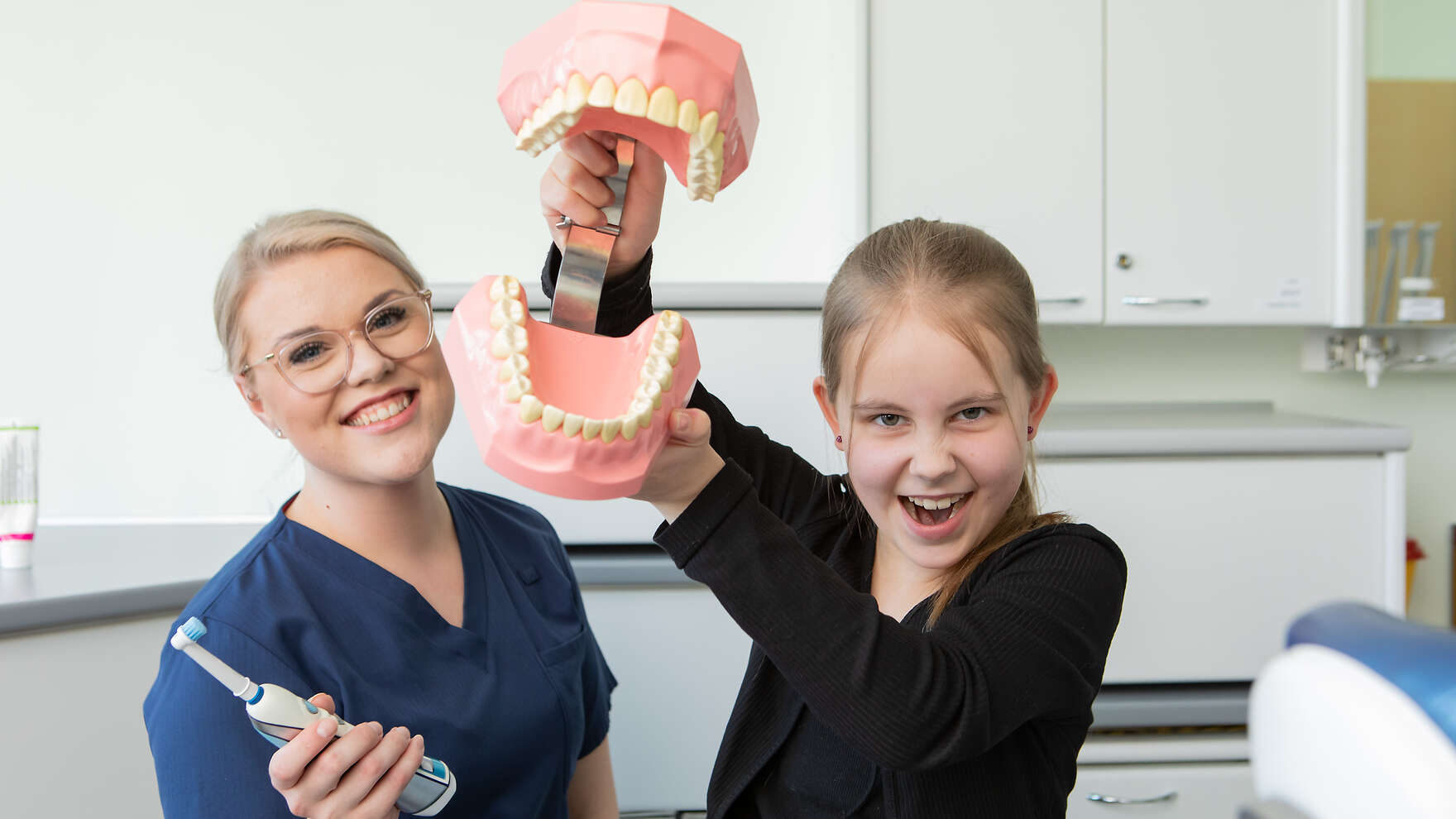 Lapsi näyttää kädessään olevia isoja tekohampaita. Vieressä on hammashoitaja, jolla on kädessään sähköhammasharja.