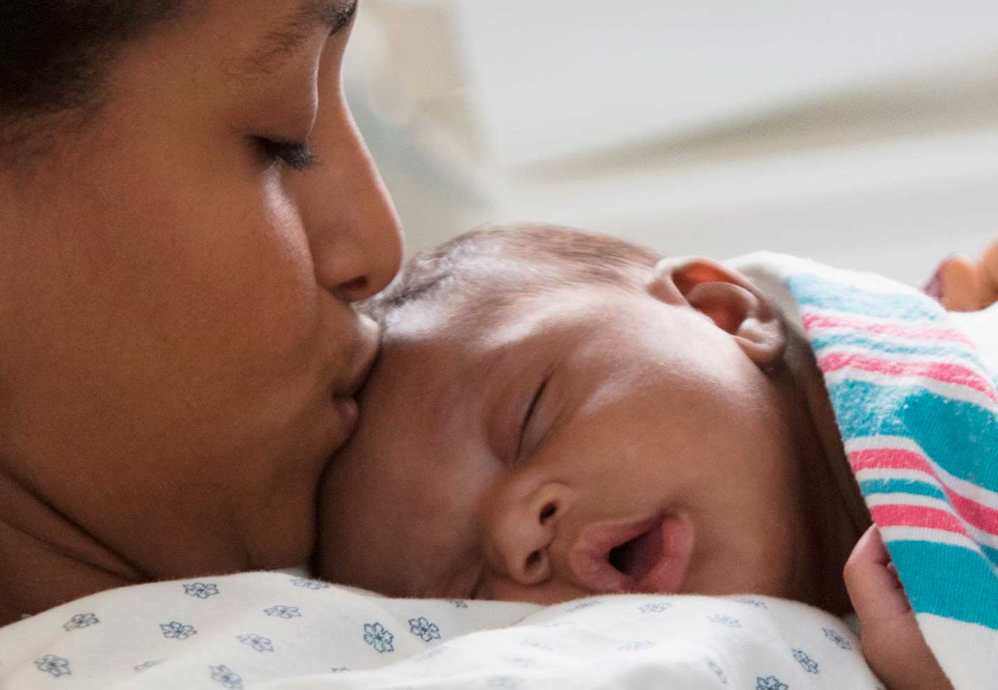 Lähikuva jossa äiti antaa pusun sylissään nukkuvan vauvan otsaan.