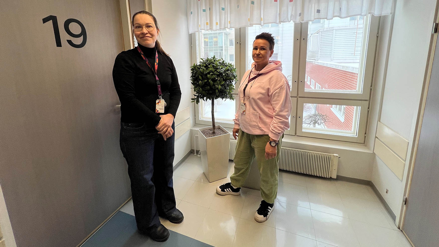 Kaksi naista seisoo käytävällä vastaanottohuoneen oven edessä, taustalla on ikkuna.