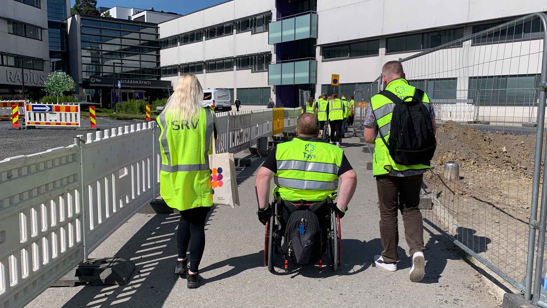 Kuvassa kaksi henkilöä kävelee ja kolmas kulkee pyörätuolilla kahden työmaa-aidan välissä kulkevaa reittiä.