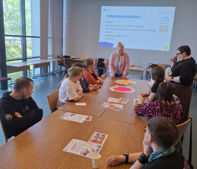 Ihmisiä on pöydän äärellä katsomassa diaesitystä. Pöydällä on papereita.