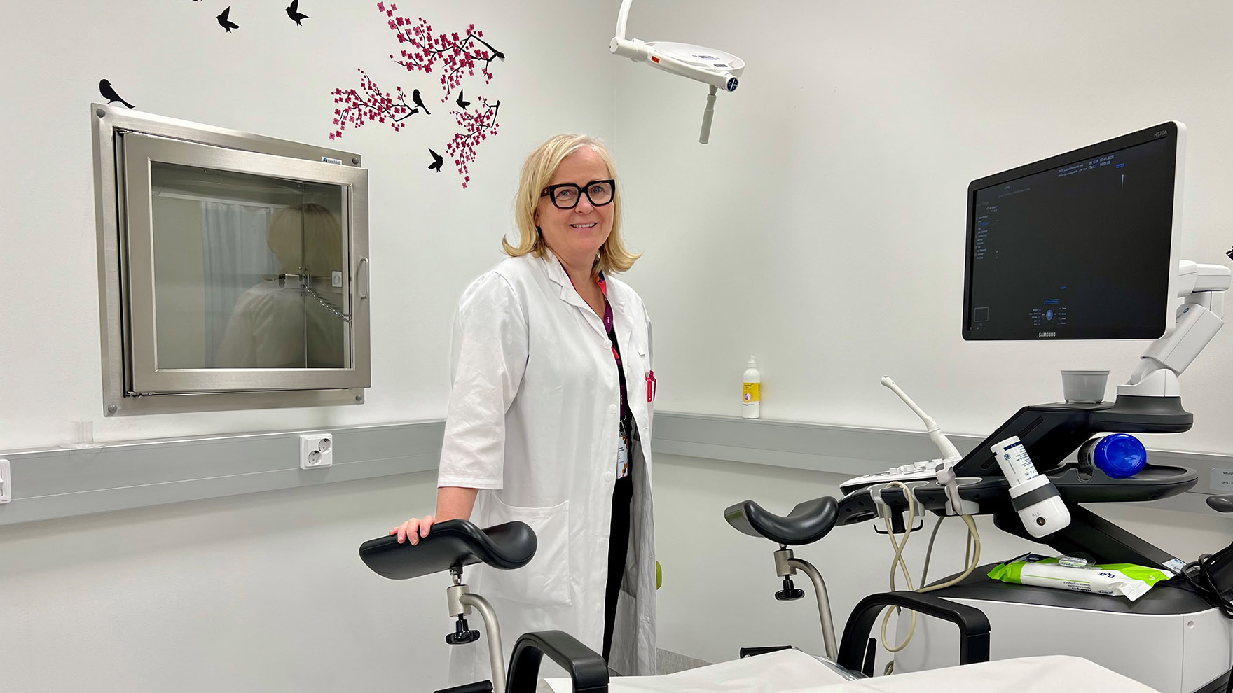 Lääkäri Katja Ahinko seisoo vastaanottohuoneessa ultraäänilaitteen vieressä ja hymyilee.