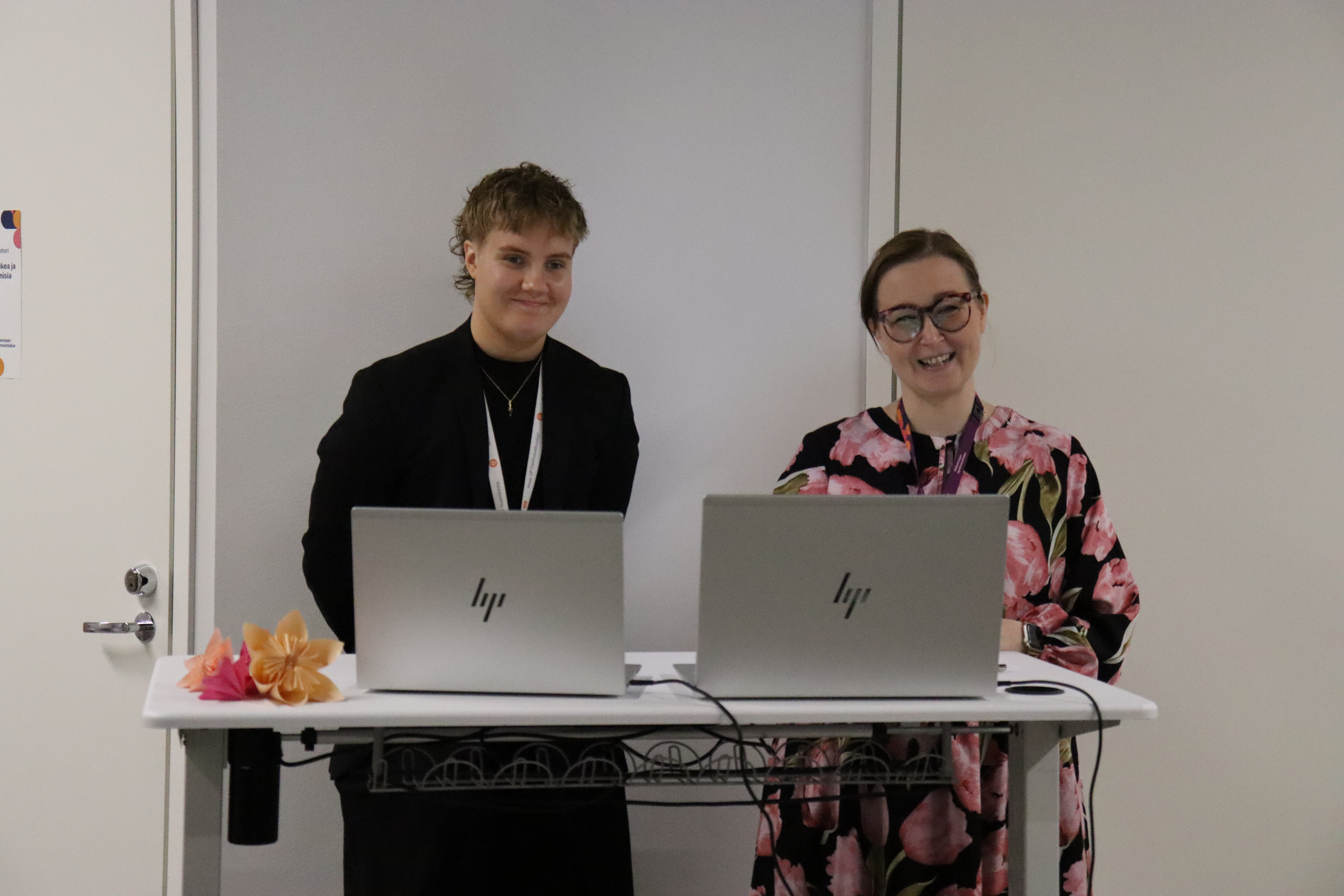 Suvi Lotvonen ja Minna Kääriäinen seisovat pöydän takana. Pöydällä on kaksi kannettavaa tietokonetta.
