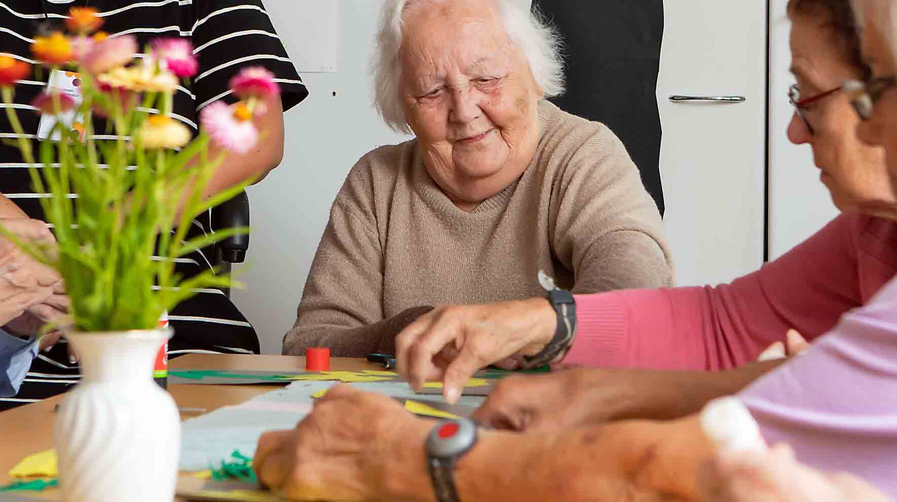 Ikääntynyt rouva istuu pöydän päässä ja hymyilee. Myös pöydän oikealla puolella istuu ikääntyneitä naisia. Pöydällä on askerteluja.