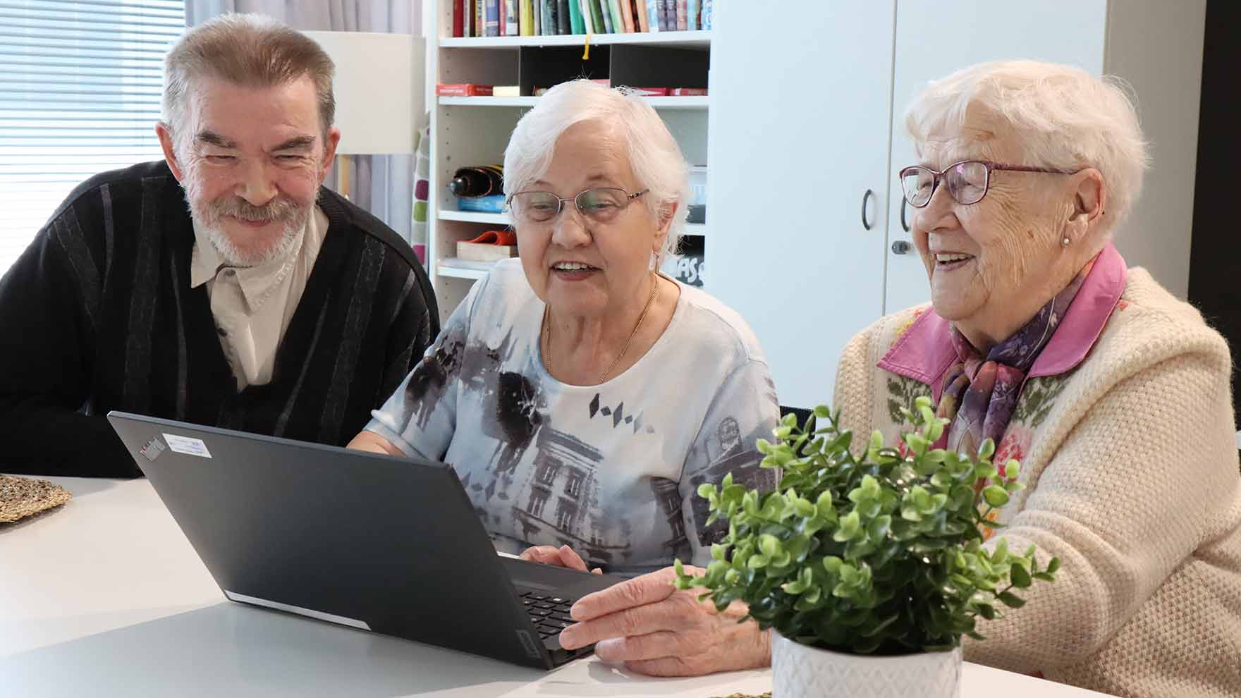 Kolme ikääntynyttä henkilö katsoo yhdessä tietokonetta hymyssä suin.