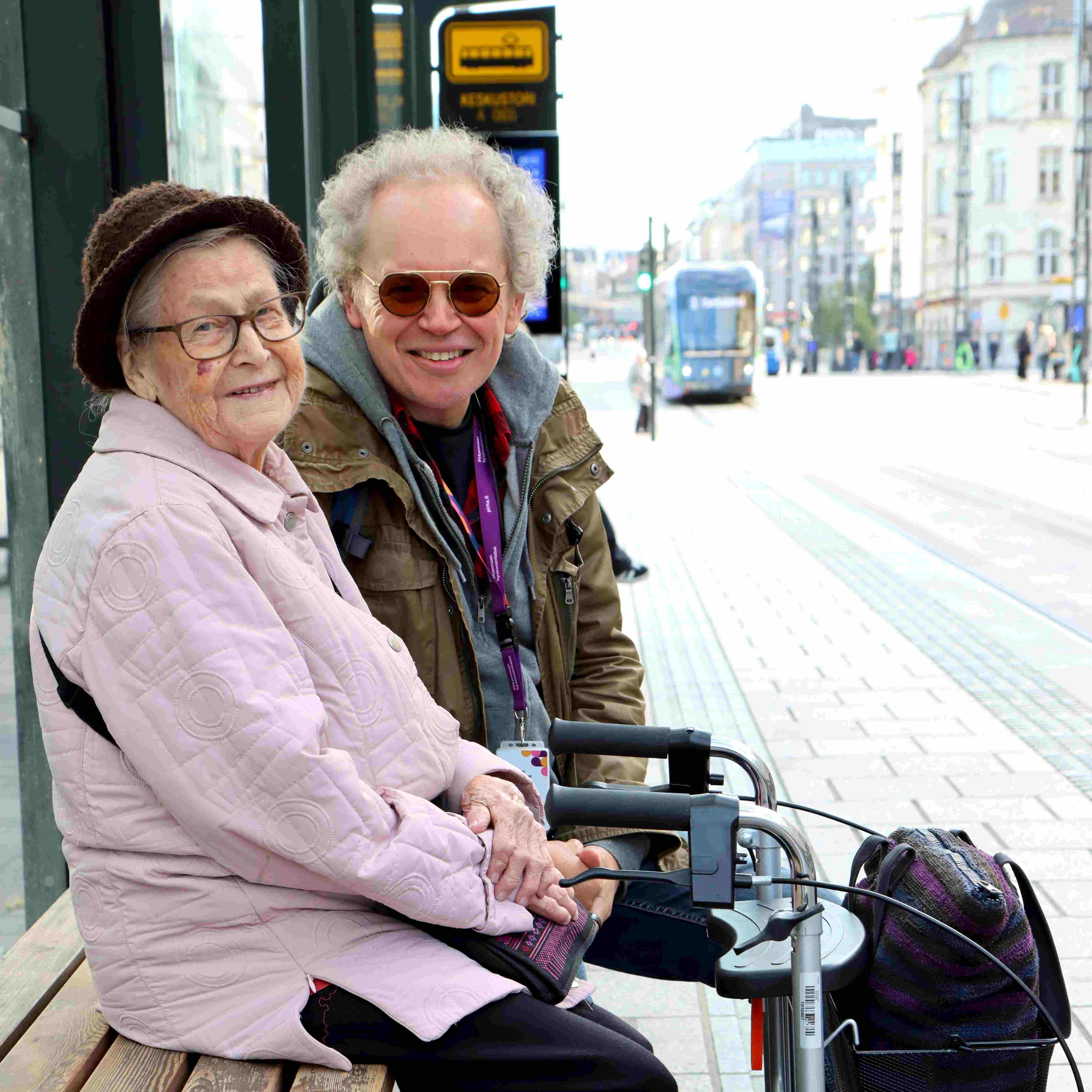 Palveluavustaja Jouni istuu ikäihmisen kanssa ratikkapysäkillä Tampereella.