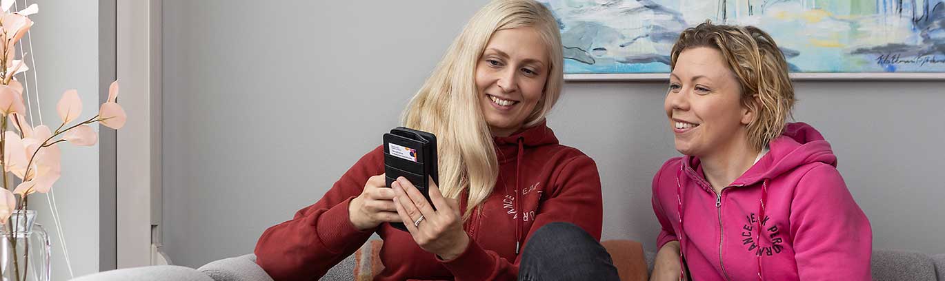 Kaksi hymyilevää naista istuu vierekkäin ja katsoo toisen kädessä olevaa kännykkää.