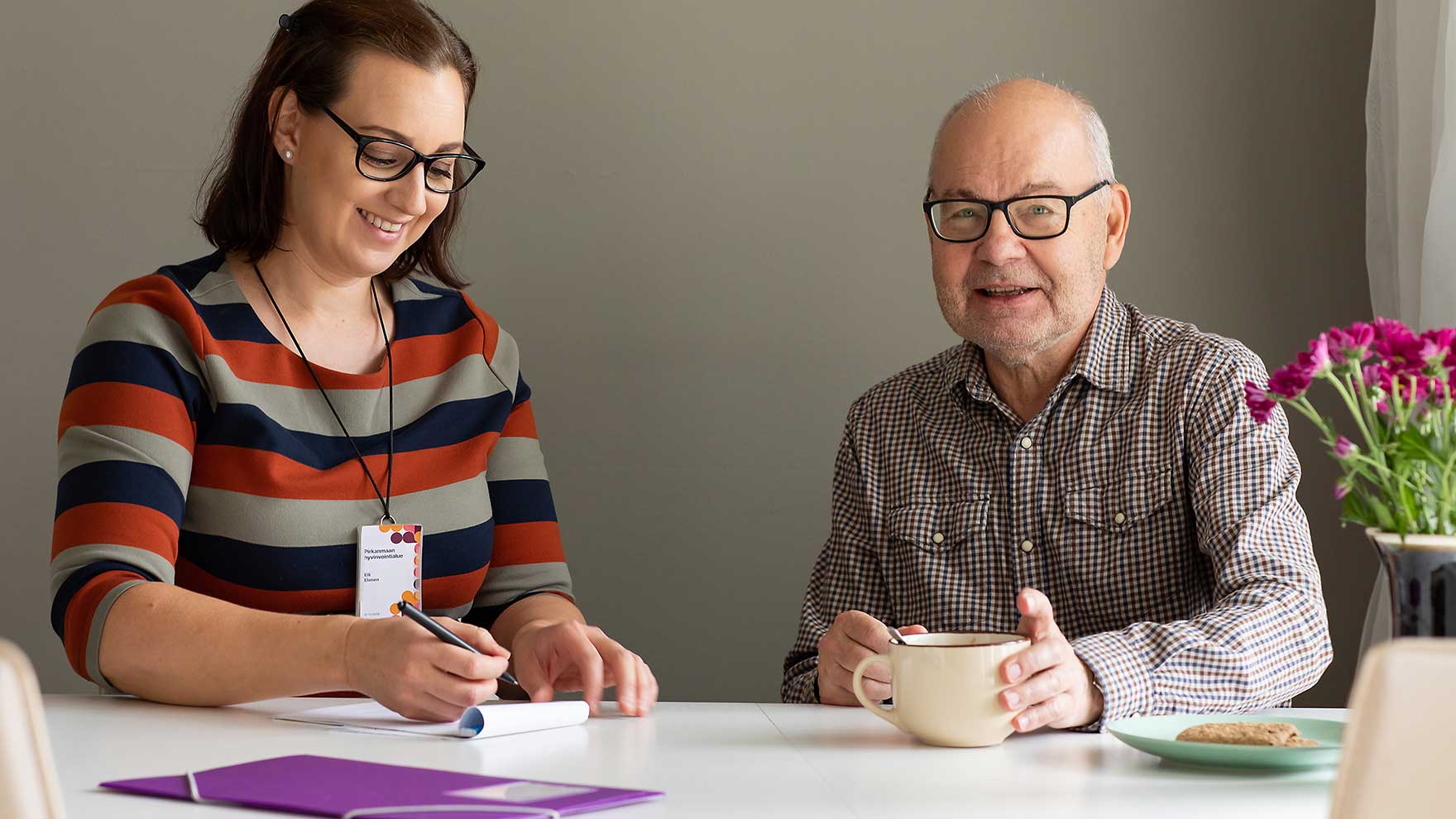 Pirkanmaan hyvinvointialueen työntekijä ja iäkkäämpi mies kahvipöydän äärellä.
