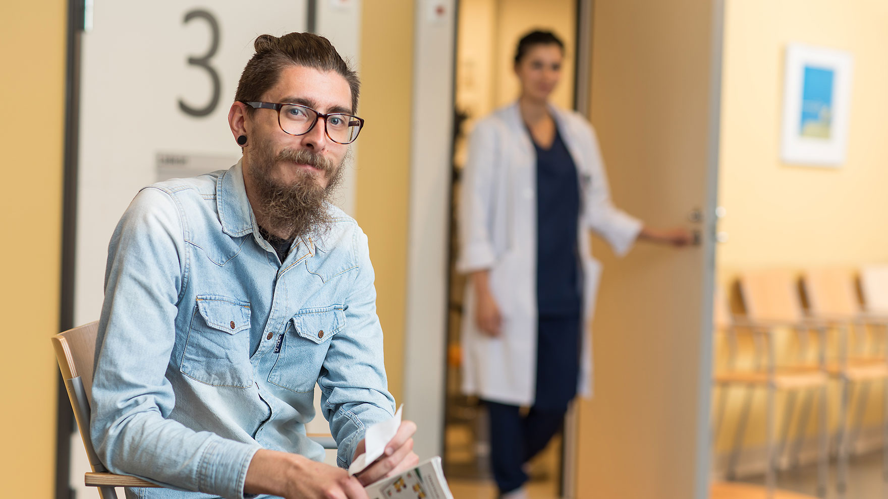 Mies istuu terveysaseman aulassa ja katsoo kohti kameraa, taustalla seisoo terveydenhuollon ammattilainen vastaanottohuoneen ovella.