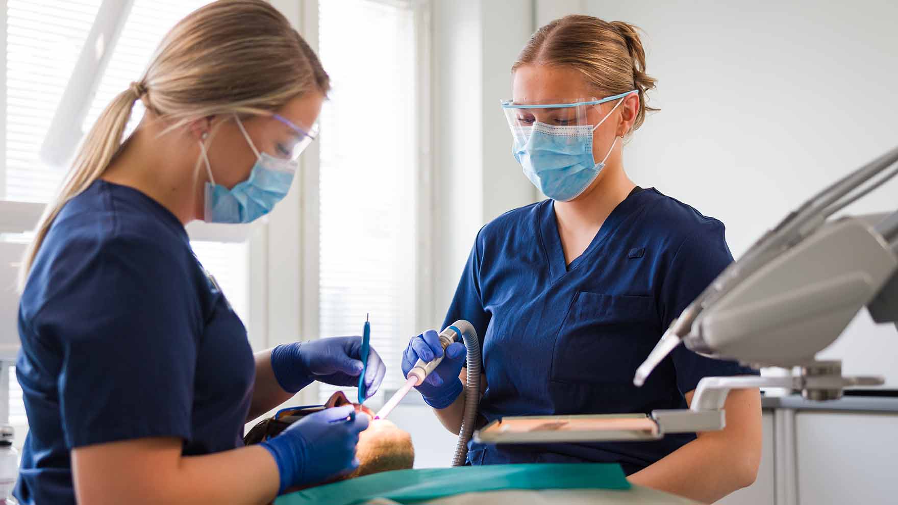 Kaksi suun terveydenhuollon ammattilaista tutkimassa makuulla olevan asiakkaan hampaita..