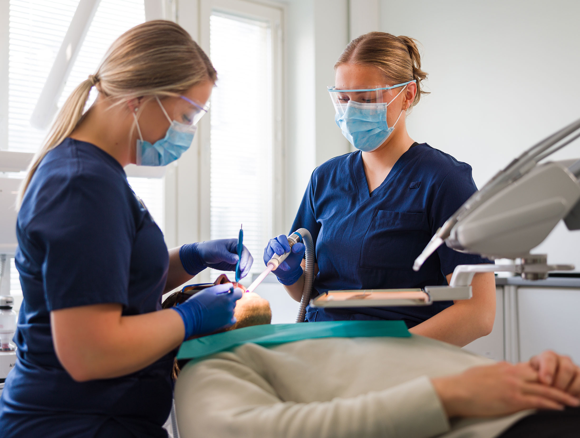 Kaksi suun terveydenhuollon ammattilaista tutkimassa makuulla olevan asiakkaan hampaita..