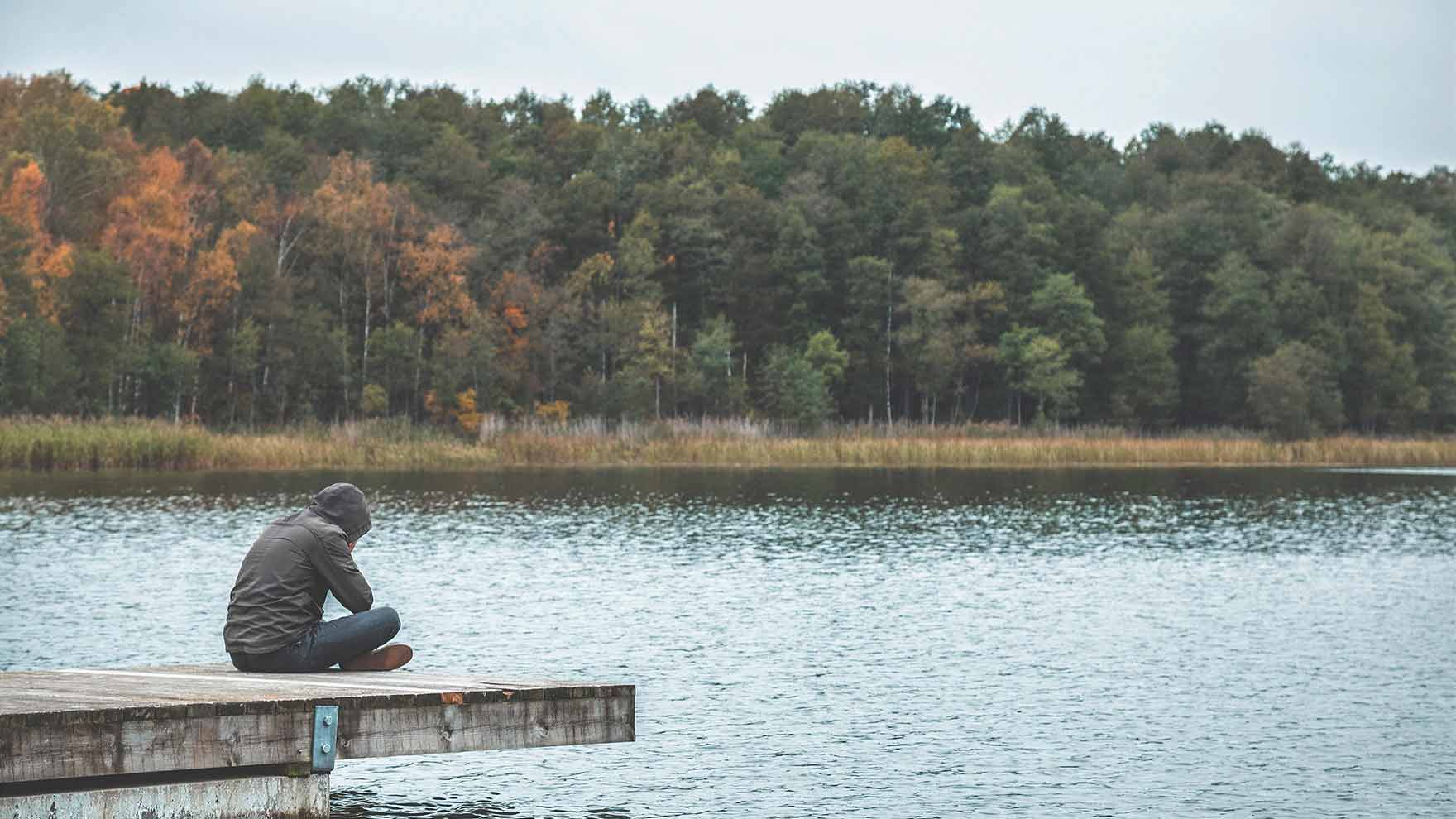 Mies istuu laiturilla huppu päässä. Järven takana näkyy syksyistä metsää.