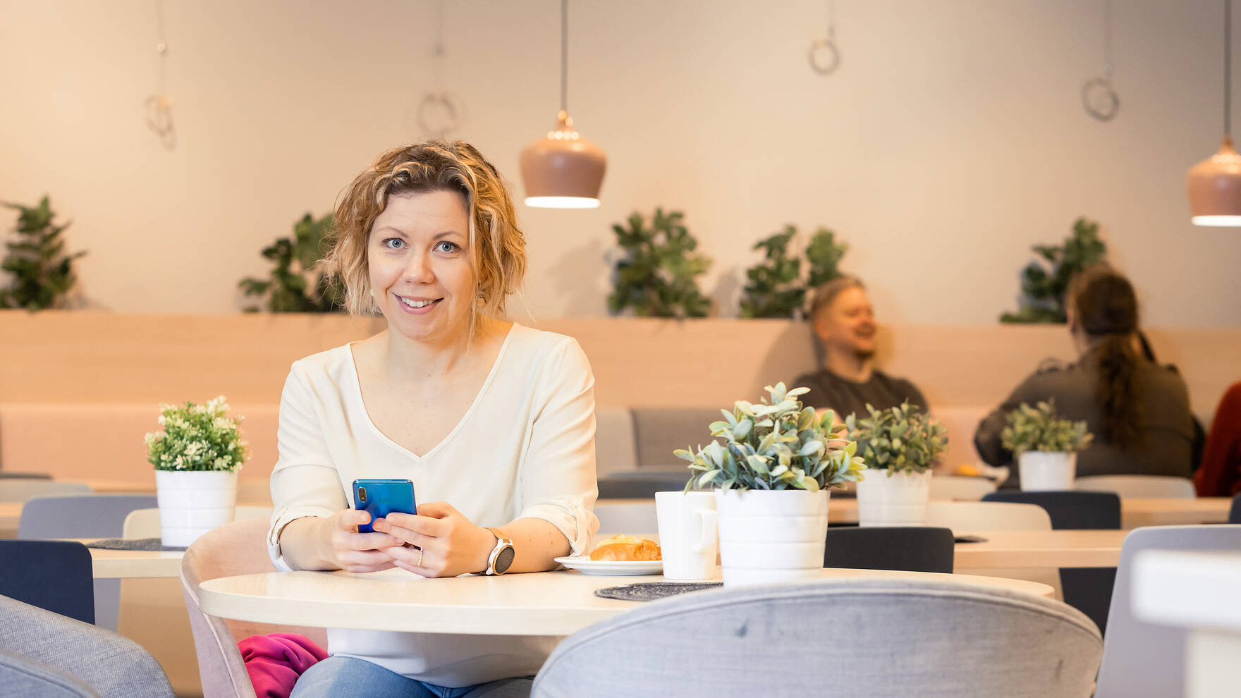 Hymyilevä, vaaleahiuksinen henkilö istuu kahvilassa pyöreän pöydän ääressä ja pitää kännykkää kädessään.