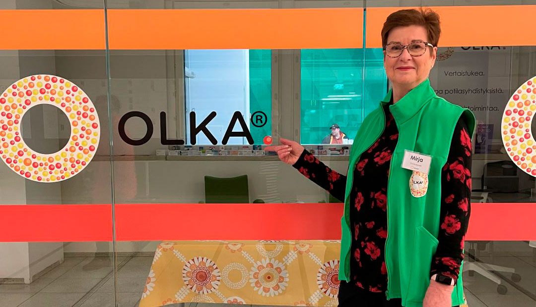 Mirja Savolainen osoittaa sormellaan OLKA-logoa lasiseinässä.