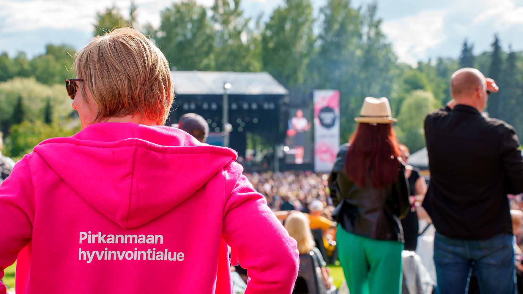 A woman is wearing a pink Wellbeing Services County of Pirkanmaa hoodie at an outdoors concert.