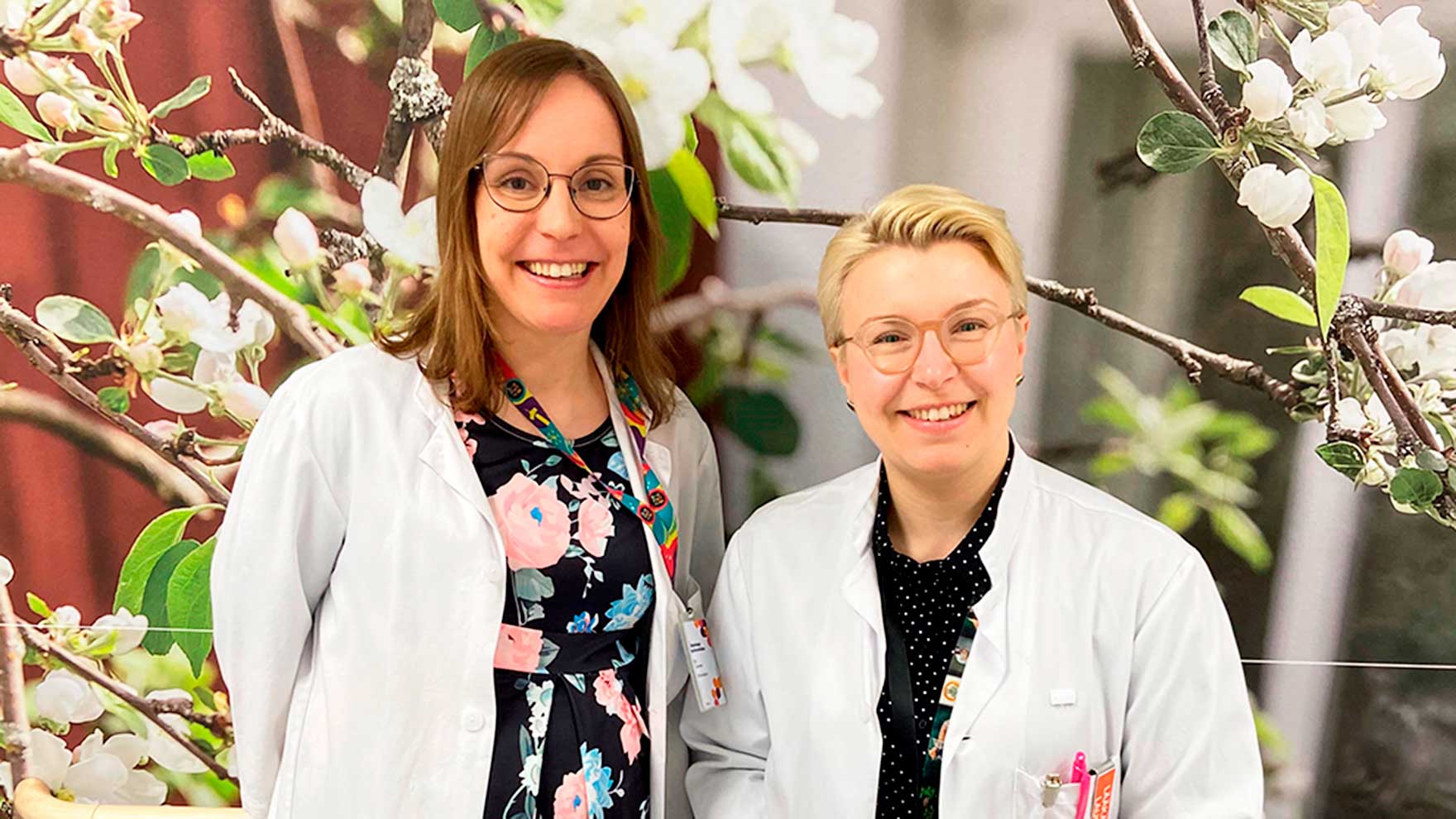 Kuvassa ovat lääkärintakki yllään Elli Toivonen ja Laura Seppälä. Taustalla näkyy valokuvatapetti, jossa on valkoisenaan kukkivia oksia.
