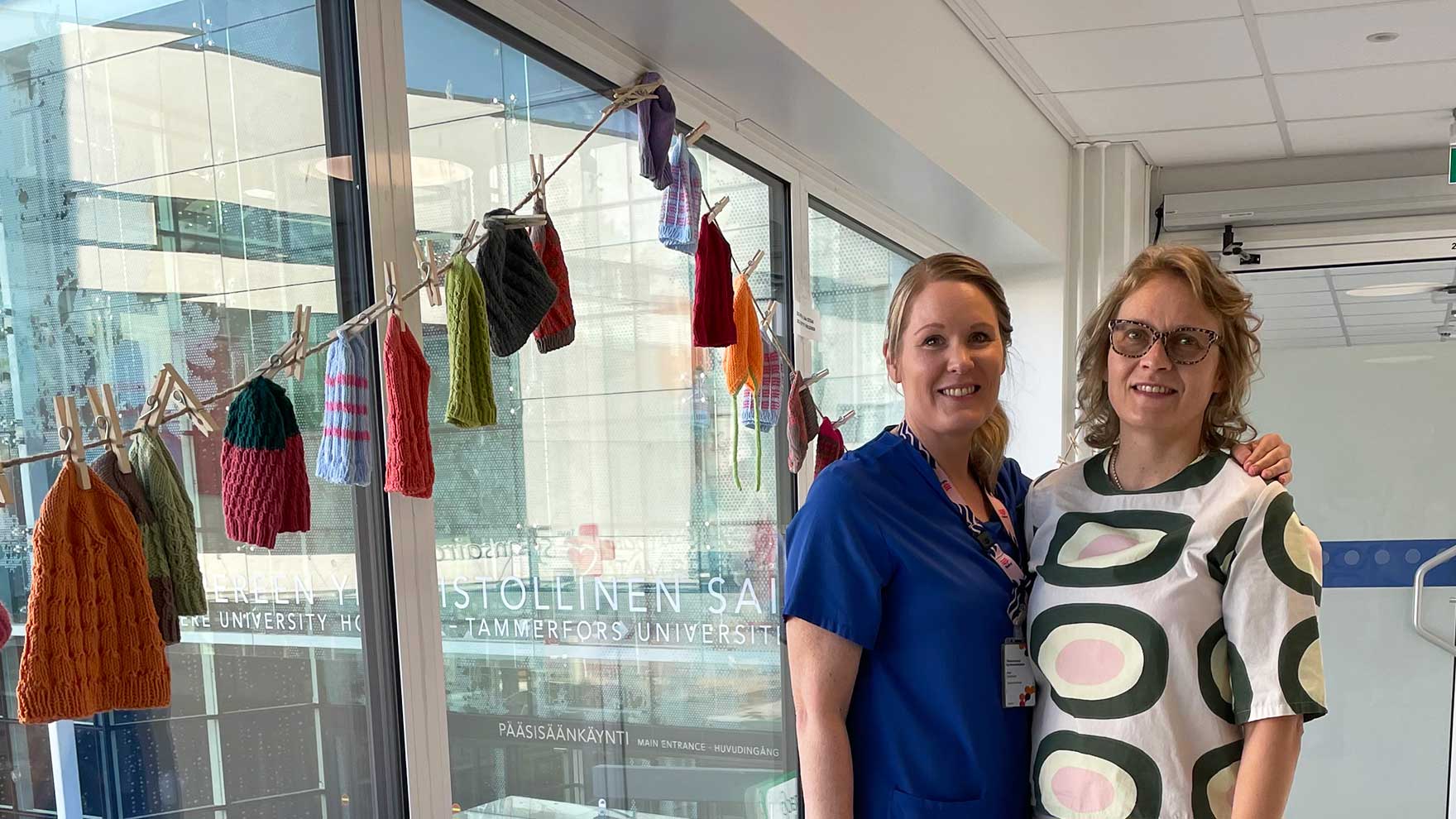 Mari Aaltonen ja Hilla Wallin seisovat yksikön aulatilassa, jonne on ripustettu neulottuja pipoja.