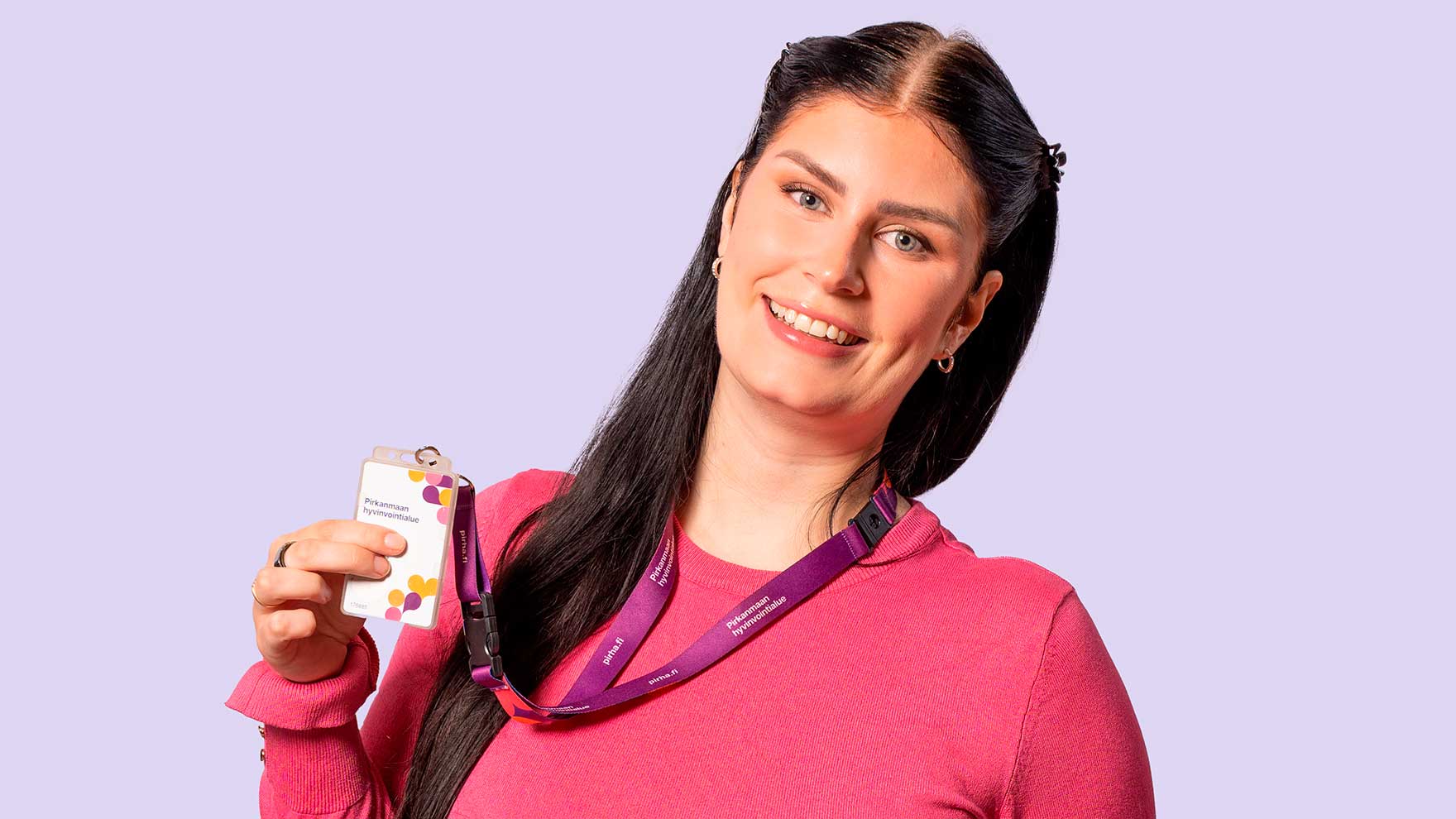 Tummahiuksinen nainen pinkissä puserossa katsoo hymyillen kohti kameraa ja pitää oikeassa kädessään esillä Pirhan henkilökorttia.
