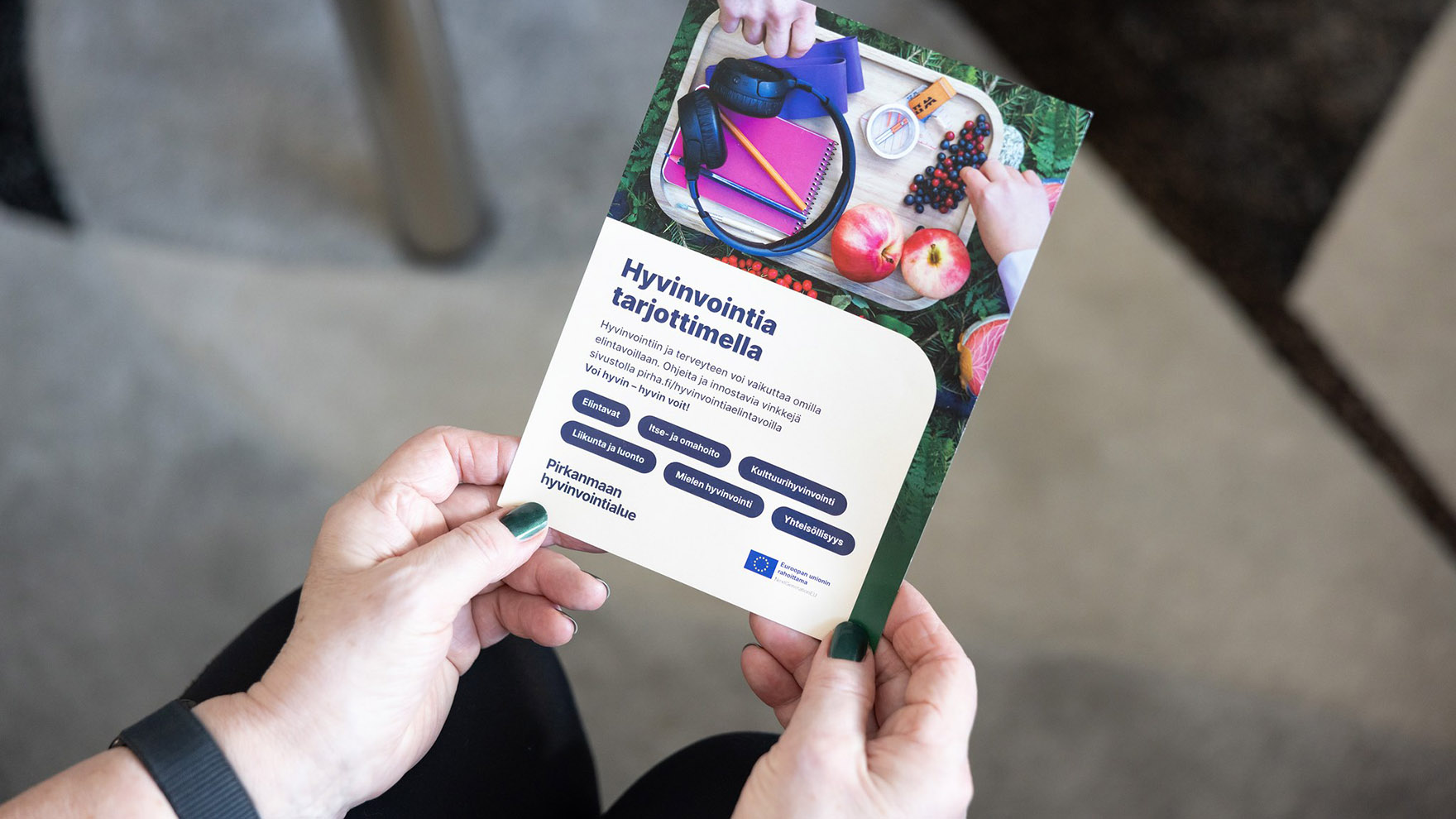 Henkilö pitää käsissään esitettä, jossa lukee ”Hyvinvointia tarjoittimella