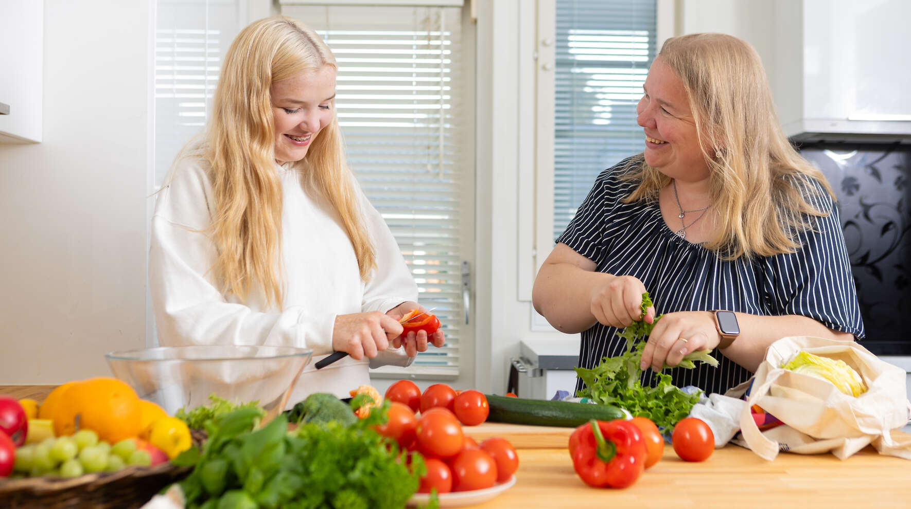 Äiti ja teini-ikäinen tyttö pilkkovat iloisina värikkäitä kasviksia.  Pöydällä on paprikoita, tomaatteja, salaattia, persiljaa, basilikaa, viinirypäleitä ja appelsiineja.