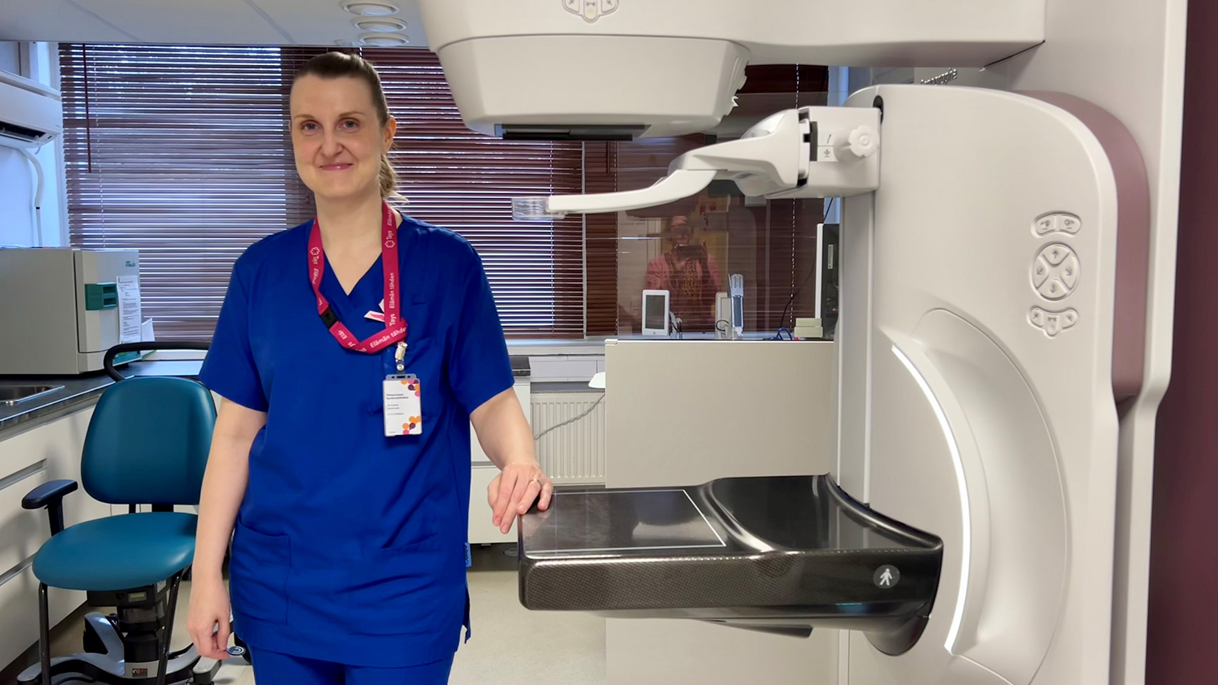 Annukka Salminen sinisissä työvaatteissaan seisoo mammografialaitteen vierellä ja hymyilee.
