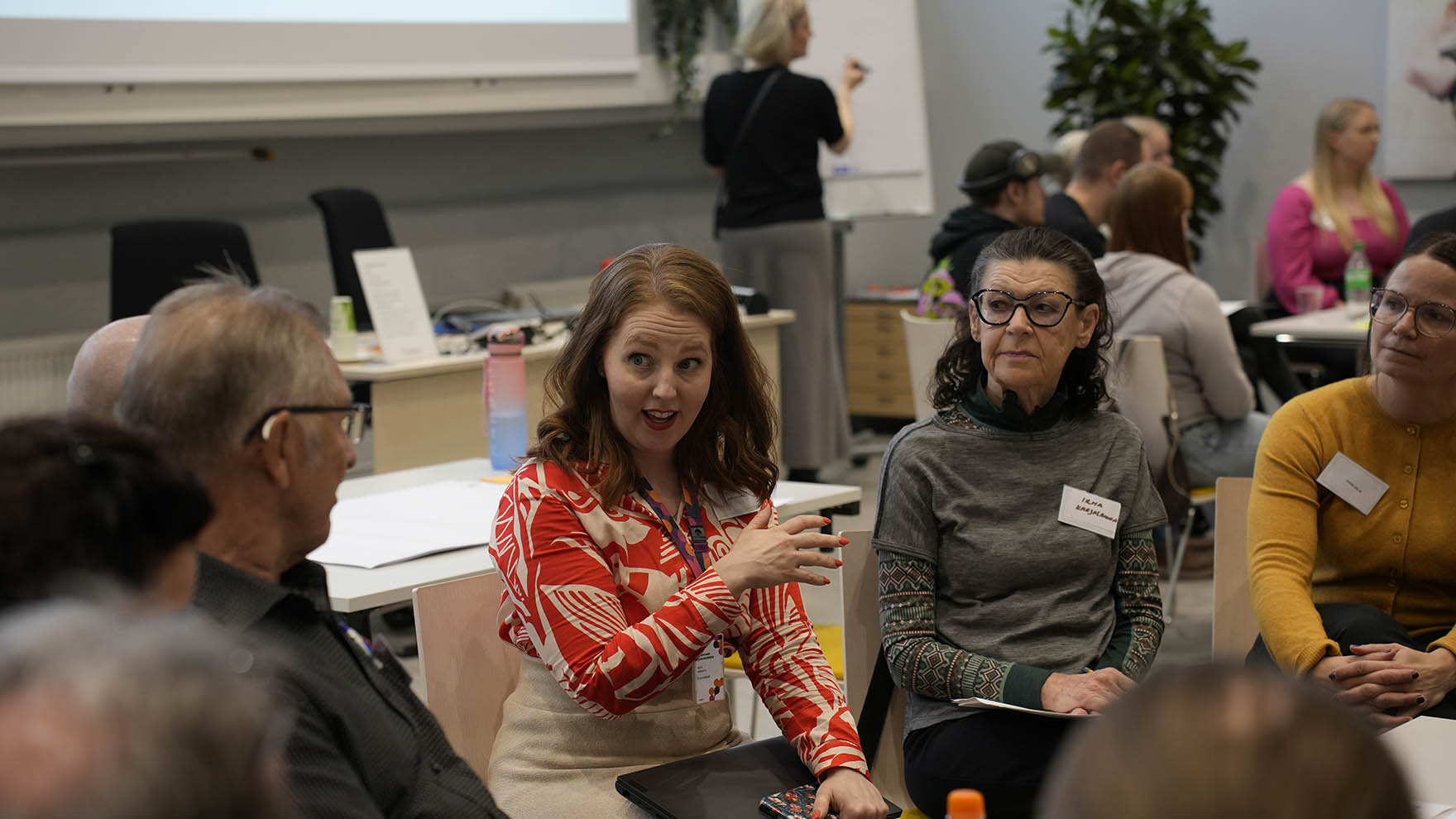 Useita ihmisiä osallistumassa asukaspaneeliin isossa salissa. Ihmiset istuvat ryhmissä ja keskustelevat keskenään.