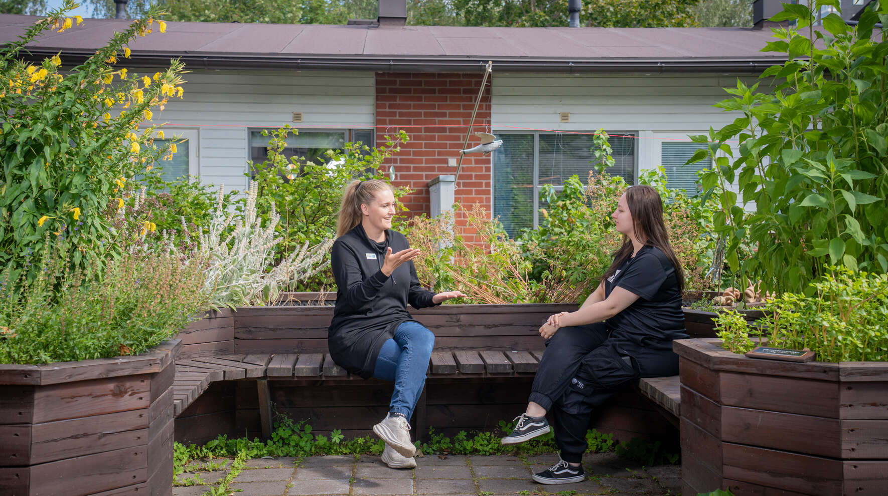 Kaksi ihmistä istuvat puutarhan penkillä, toinen viittoo käsillään toiselle.