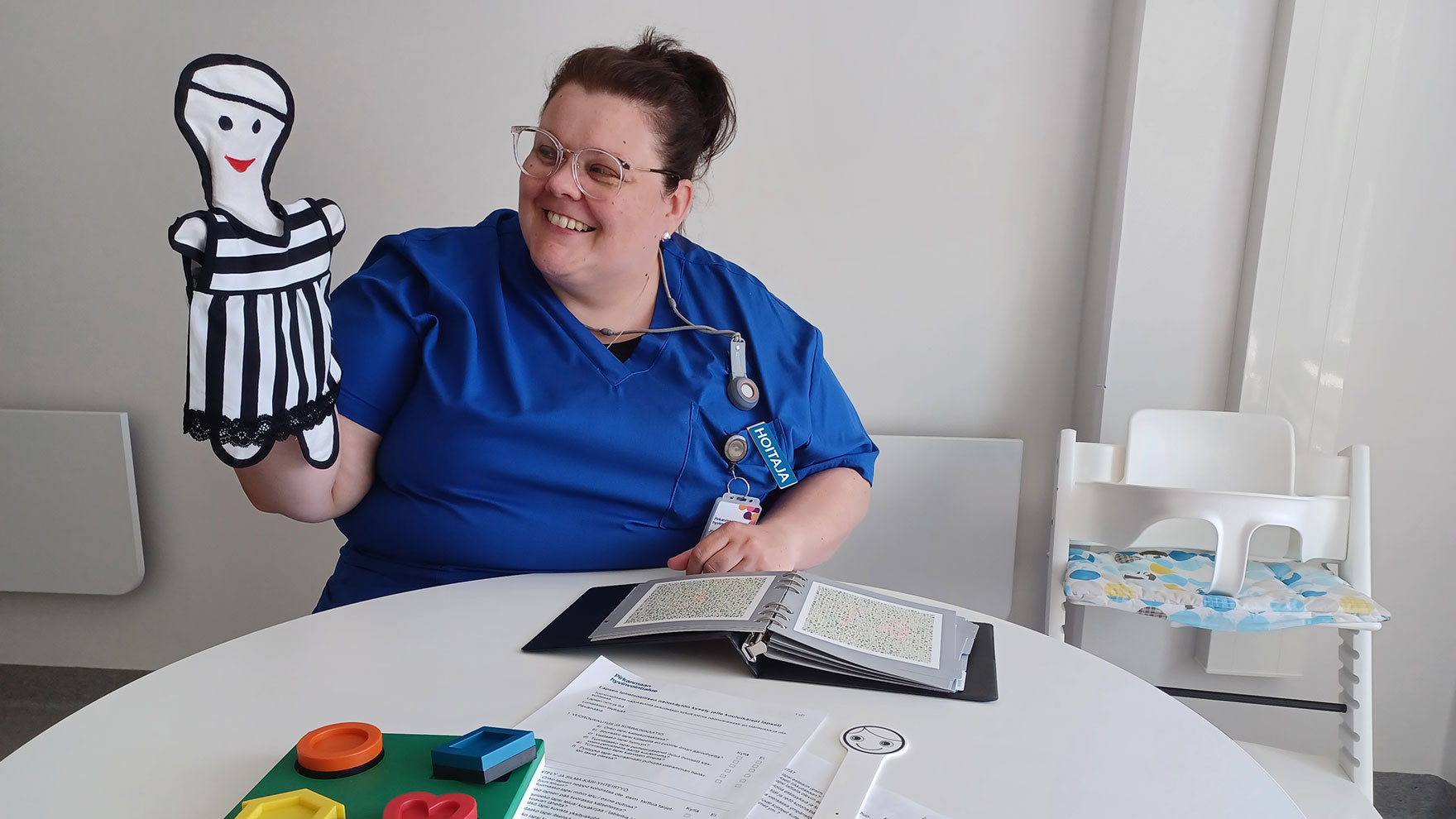 Karoliina Kilponen käsinukke kädessään. Pöydällä kansio, papereita ja peli, jolla voi harjoitella tai testata muotojen hahmottamista.