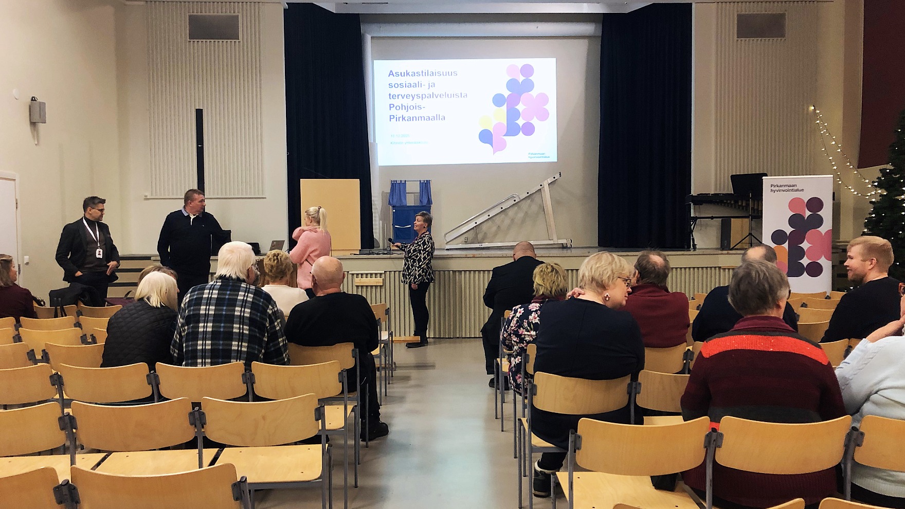 Koulun juhlasalissa ihmisiä osallistumassa asukastilaisuuteen