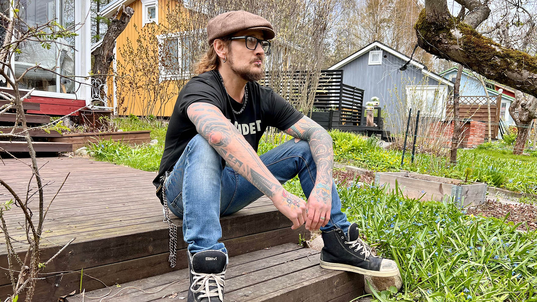 Mies istuu ulkona rappusilla. Hänellä on silmälasit ja lakki päässä.