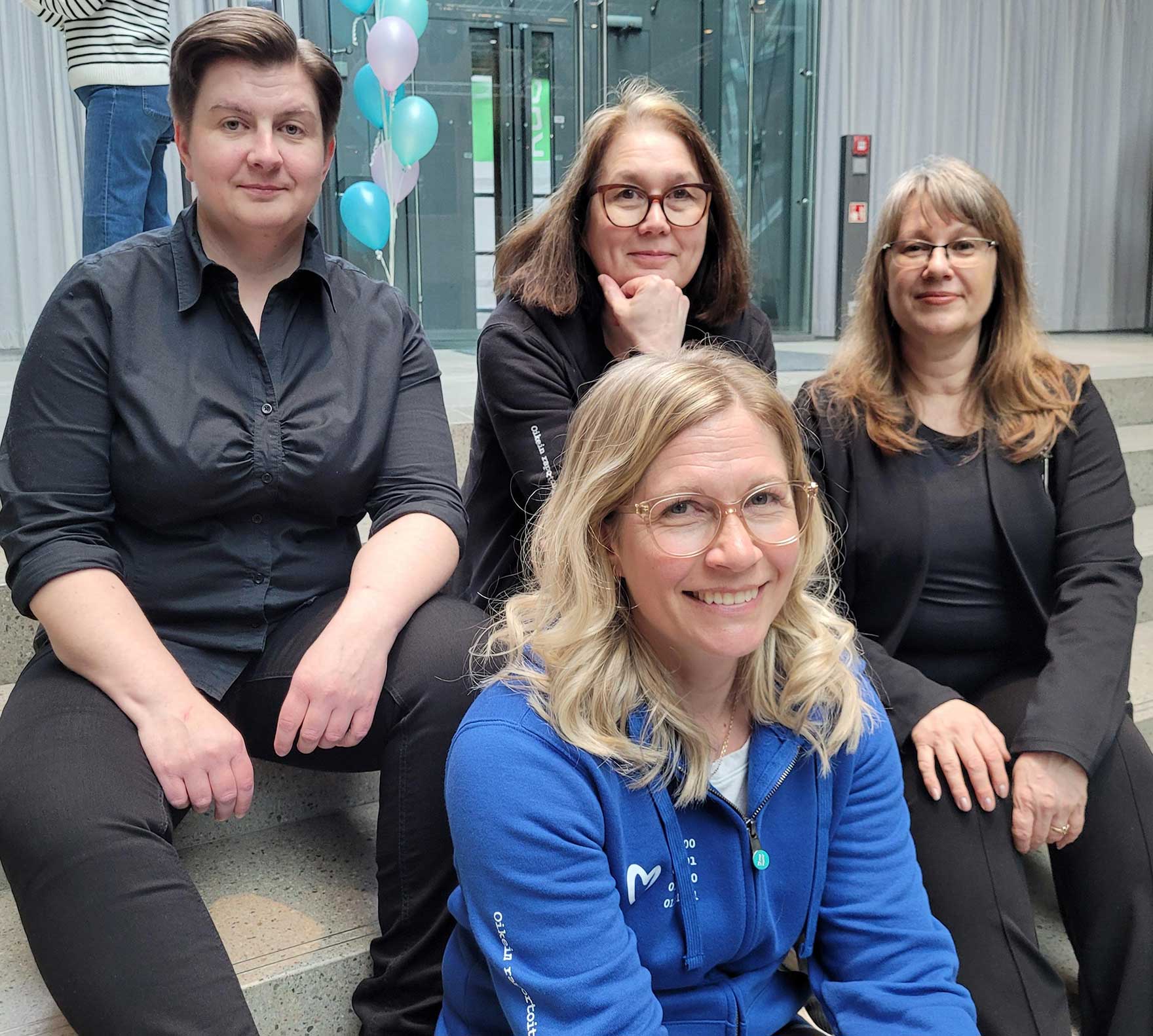 Neljä naista istuu portailla ja katsoo hymyillen kameraan.