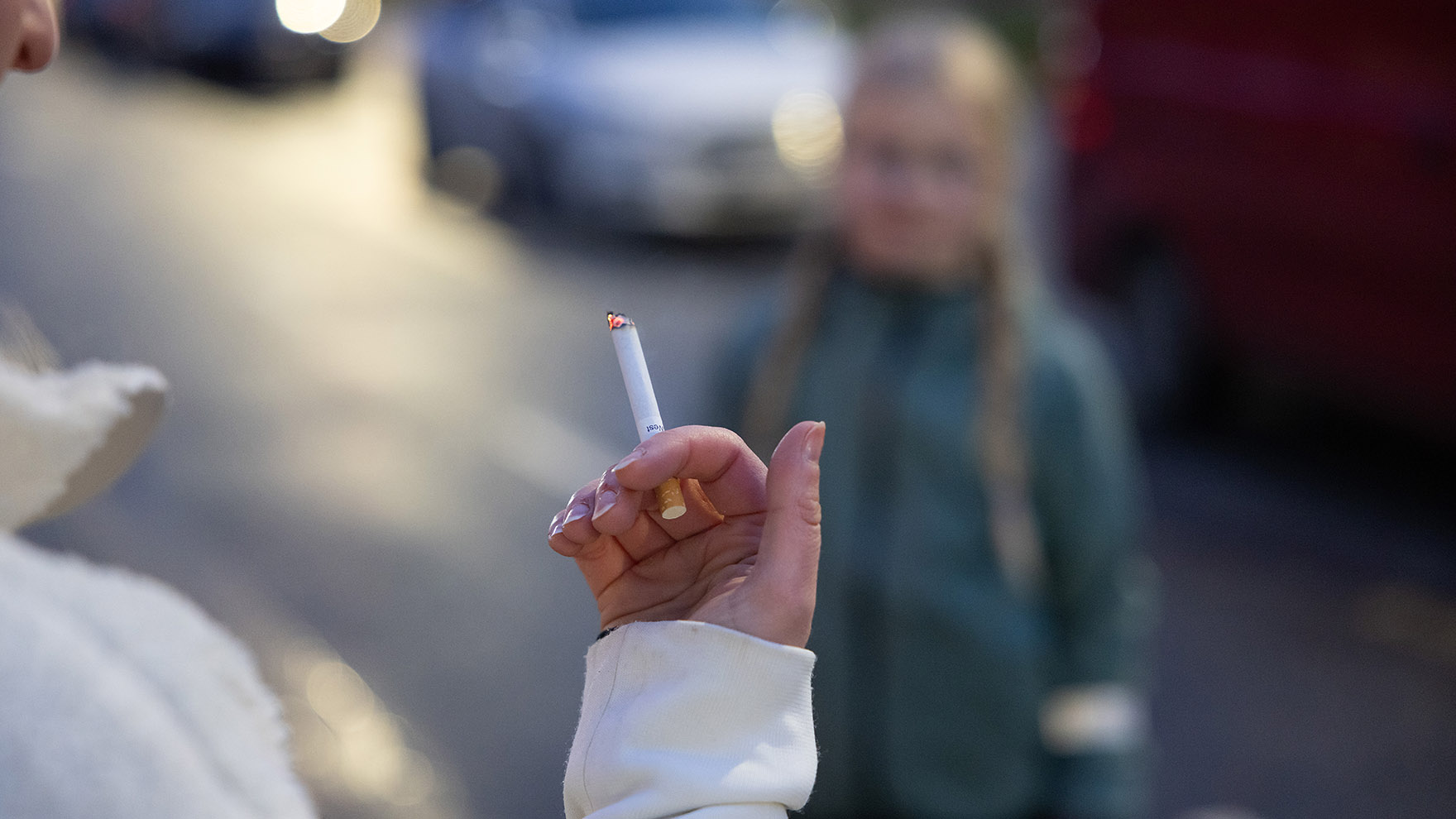 Ihminen selin tupakka kädessä ulkona. Taustalla nuori ihminen katsomassa.