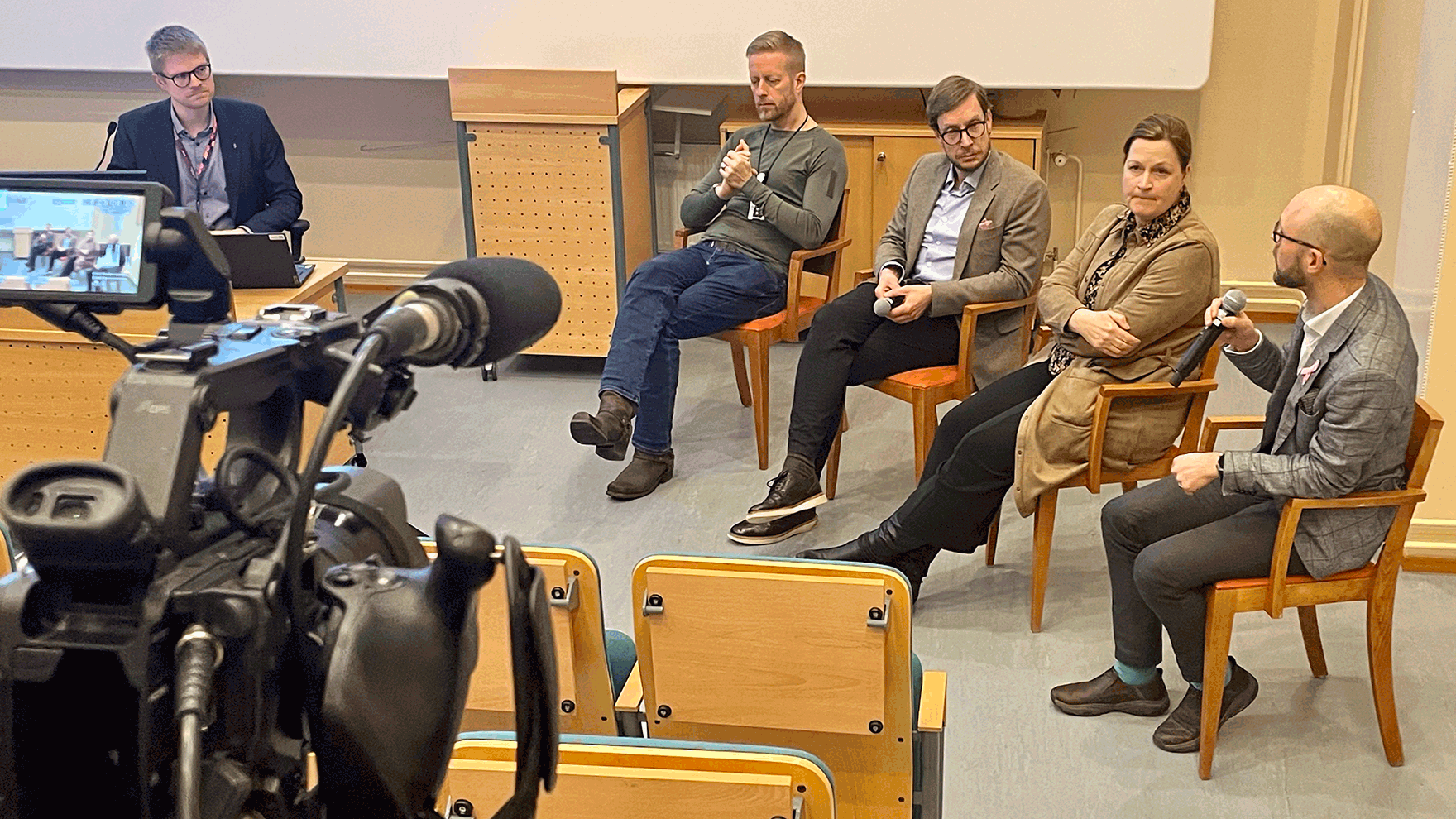 Neljä panelistia ja puheenjohtaja istuvat ja keskustelevat. Etualalla videokamera.