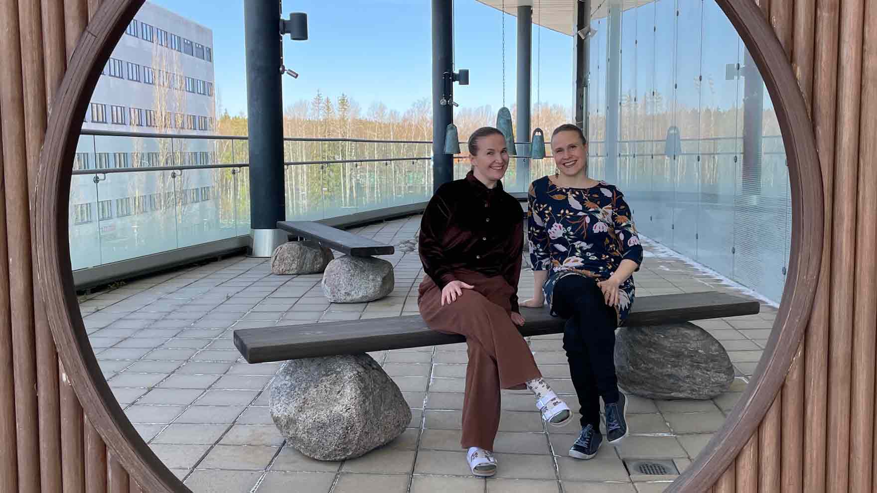 Kulttuuriohjaajat Elina ja Hanna istuvat kivistä ja laudasta tehdyllä penkillä avarassa tilassa, josta näkyy ikkunoista maisemaa.
