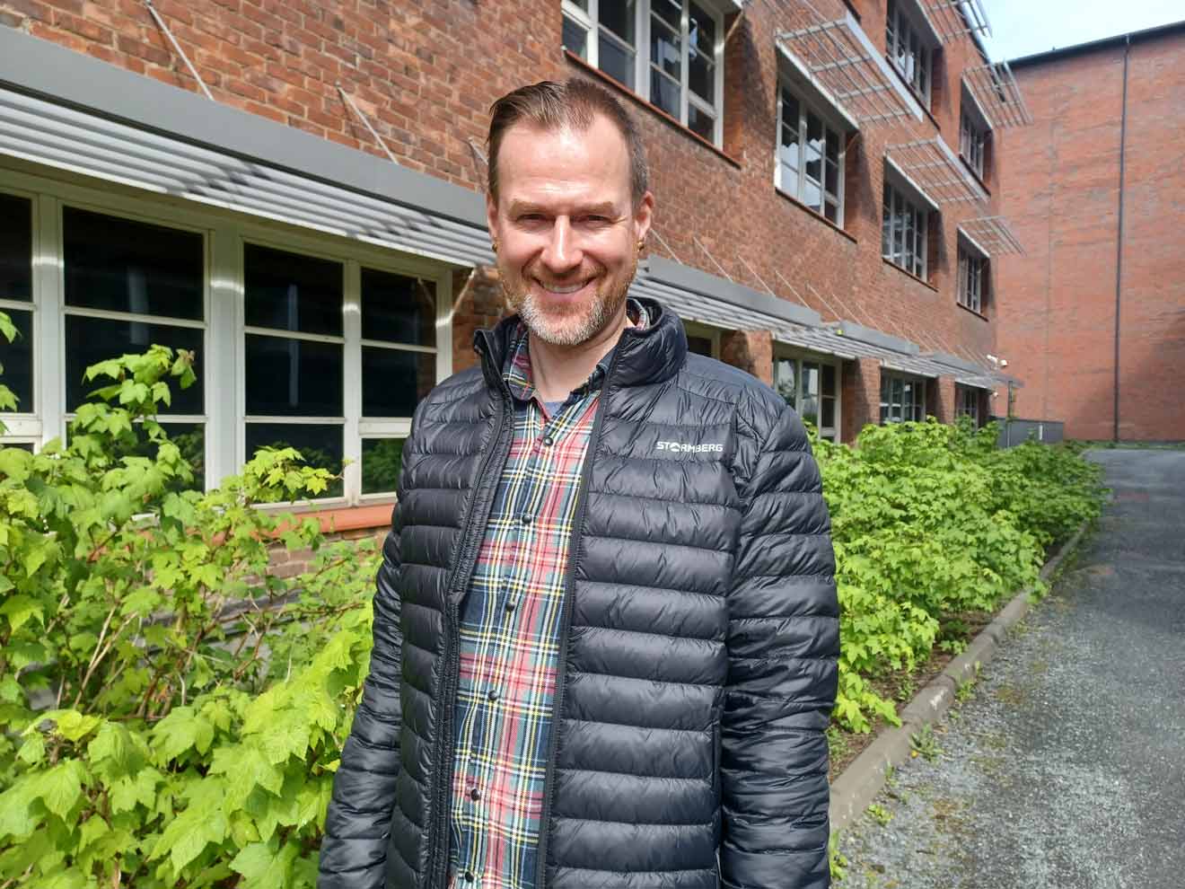 Etsivän työn ohjaaja Harri Meskanen hymyilee tiilirakennuksen ja vihreiden pensaiden edessä.