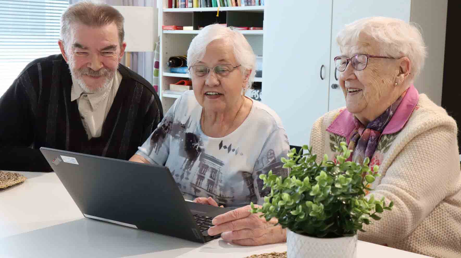Ikääntynyt herra ja kaksi rouvaa katsovat kannettavan tietokoneen näyttöä hymyillen.