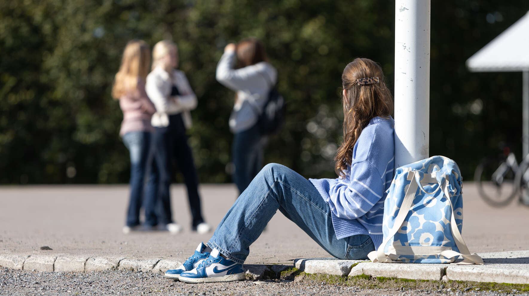 Nuori istuu yksin koulun pihalla katsellen taustalla olevaa ryhmää.