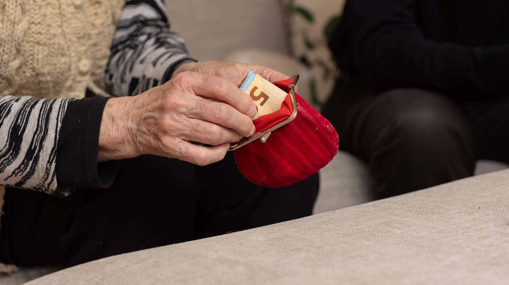 Ikääntynyt henkilö istuu sohvalla ja ottaa punaisesta kukkarosta seteleitä.