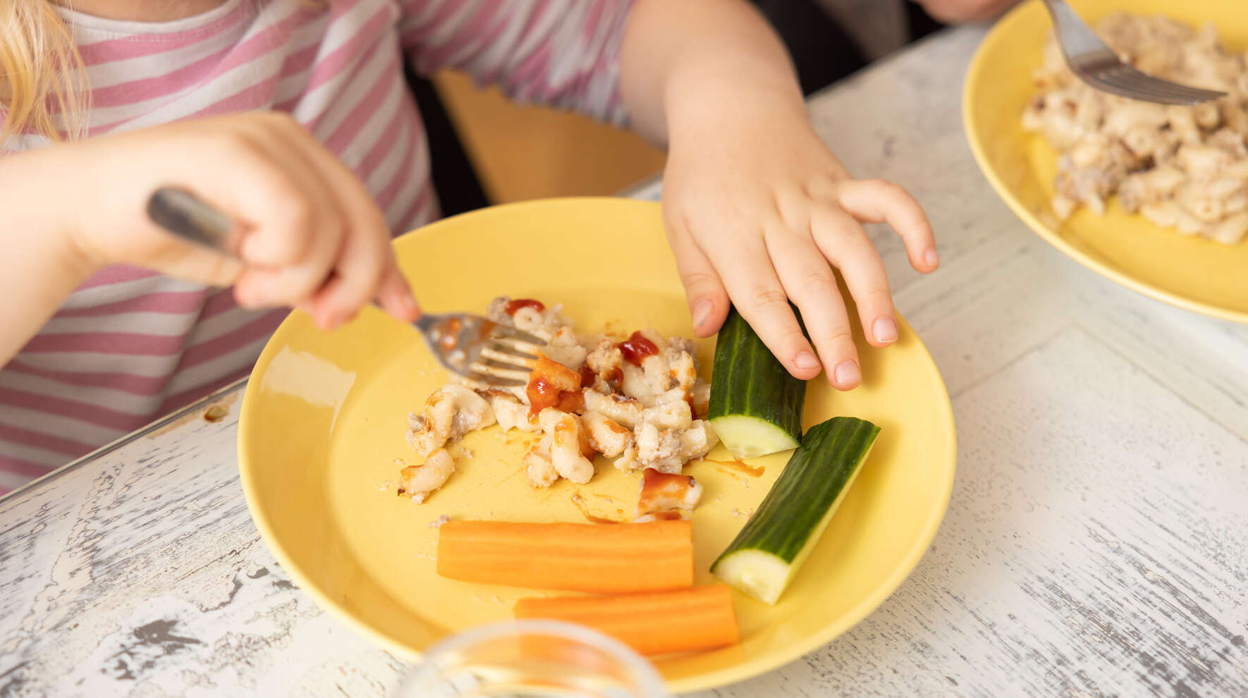 Ruokapöydällä keltainen lautanen, jossa makaronilaatikkoa ketsupilla sekä porkkana-ja kurkkutikkuja. Ruokaa syövästä lapsesta näkyy kädet.