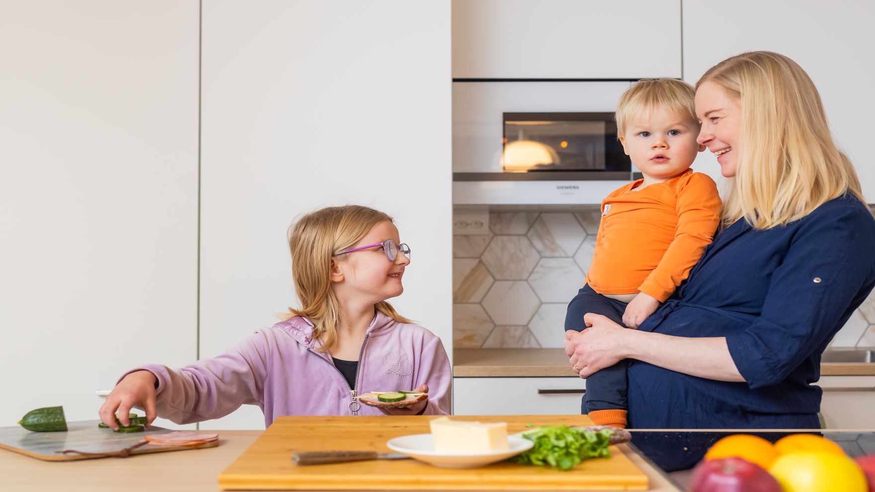 Alakouluikäinen lapsi valmistee itselleen voileipää työtason ääressä ja raskaana oleva äiti pienempi lapsi sylissään katselee hymyillen vieressä.