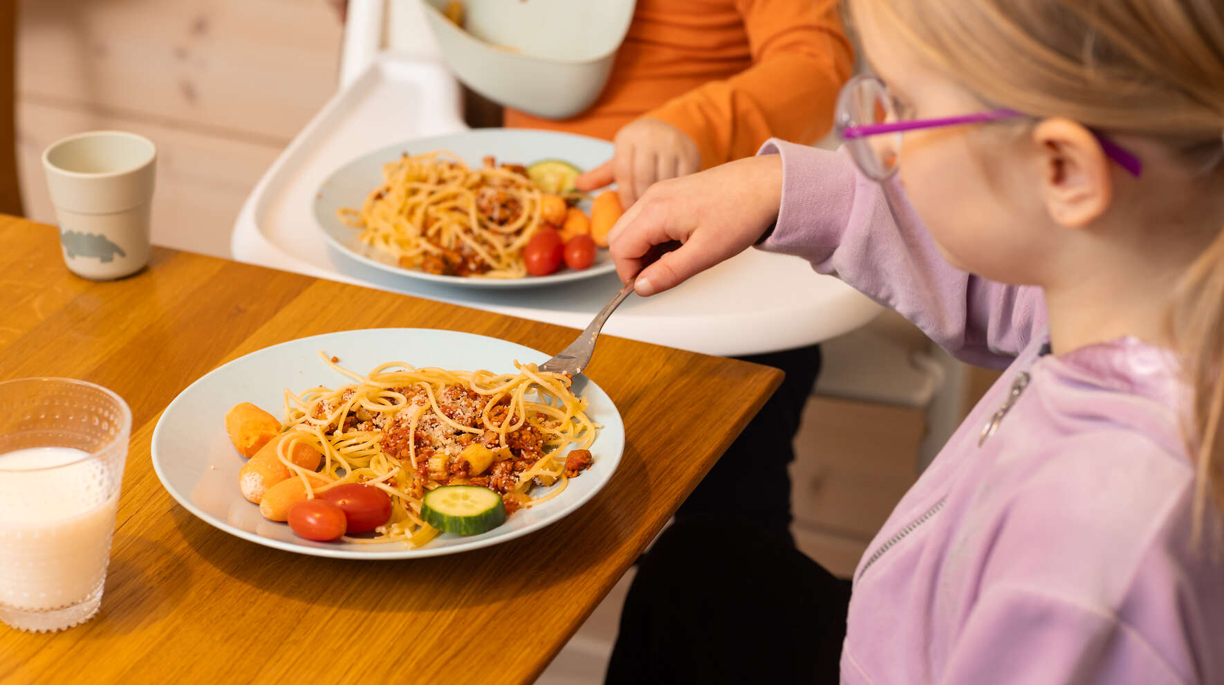 Kuvassa on kaksi lapsen ruokalautasta. Toinen on ruokapöydällä, toinen syöttötuolin tarjottimella. Lapset näkyvät vain osittain.