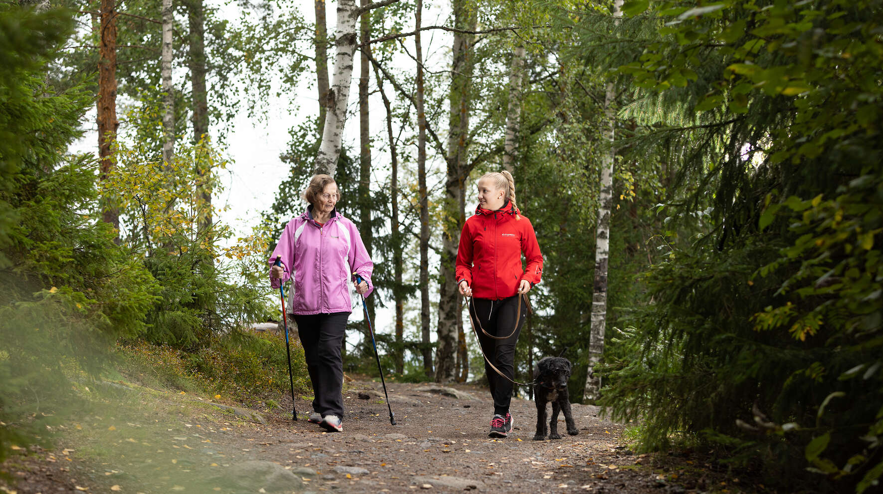 Ikääntynyt nainen sauvakävelee metsäpolulla yhdessä nuorempi naisen kanssa, jolla on koira mukana.