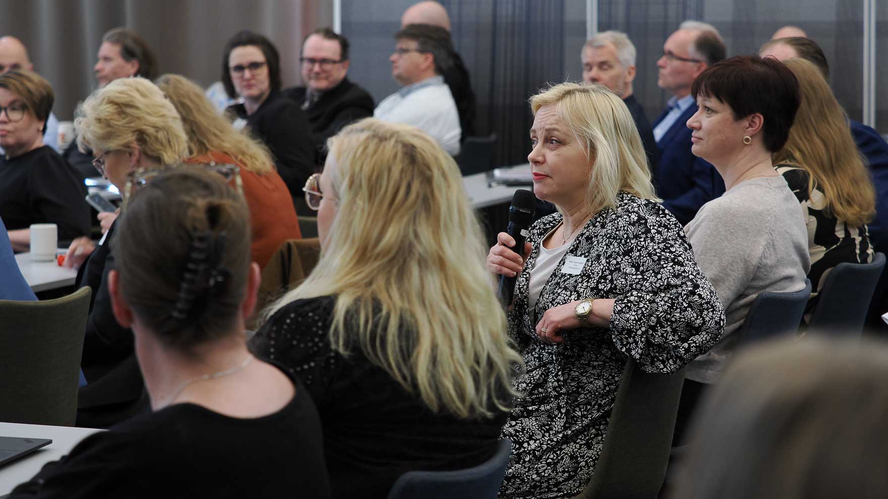 Nainen puhuu mikrofoniin seminaarisalissa. Ympärillä olevat ihmiset kuuntelevat.