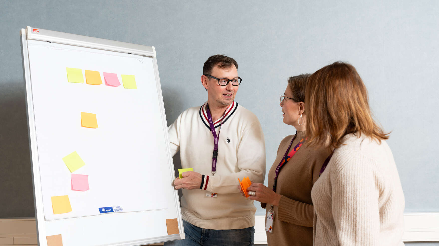 Mies ja kaksi naista seisovat flap-taulun vieressä ja laittavat siihen eri värisin post it-lappuja.