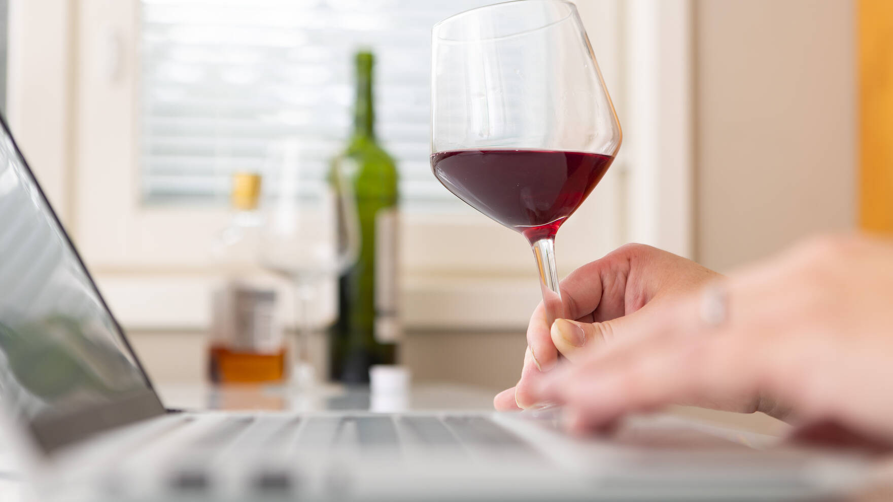 Henkilö istuu pöydässä läppärin ääressä viinilasi kädessä ja taustalla näkyy alkoholijuomapulloja ja toinen lasi.