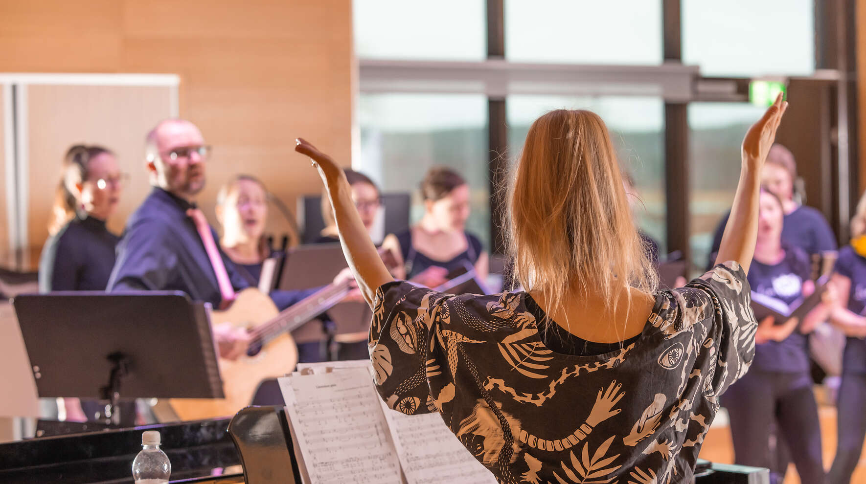 Kuoronjohtaja kädet ylhäällä selin kameraan ja taustalla näkyy kuorolaisia.