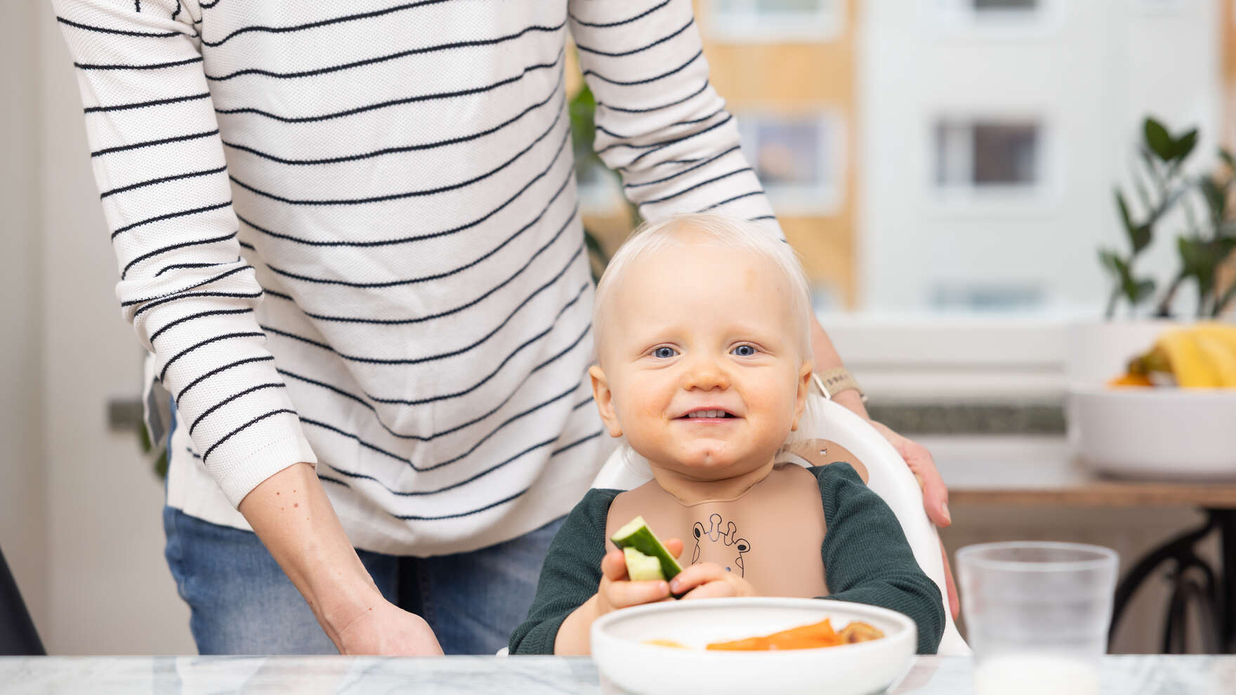 Taaperoikäinen istuu ruokapöydässä lautanen edessään ja pitää kurkkutikkuja kädessään. Taustalla näkyy vanhempi.